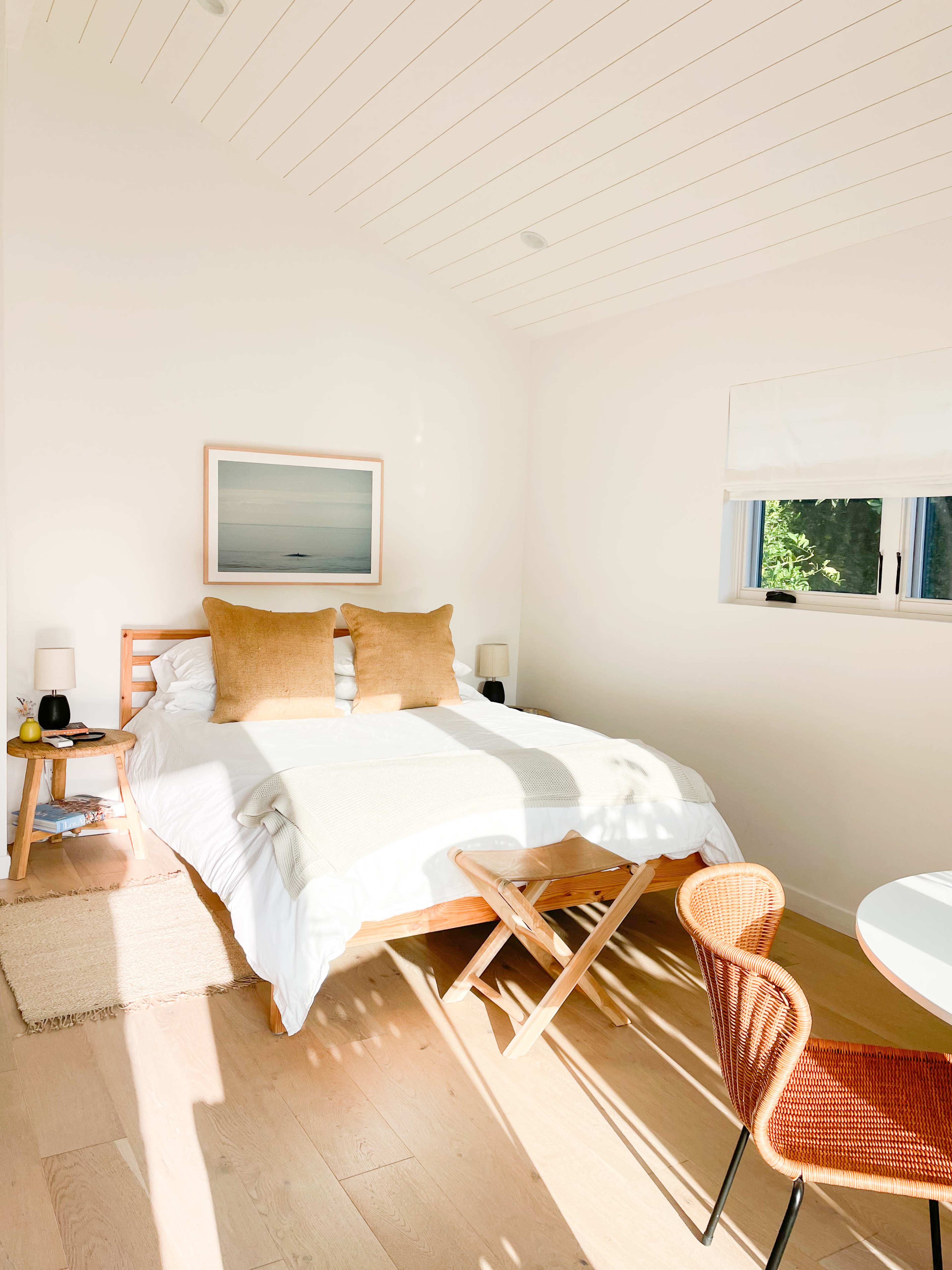 A cozy bedroom with a bed featuring beige pillows, a small side table, and a wicker chair next to a round table.