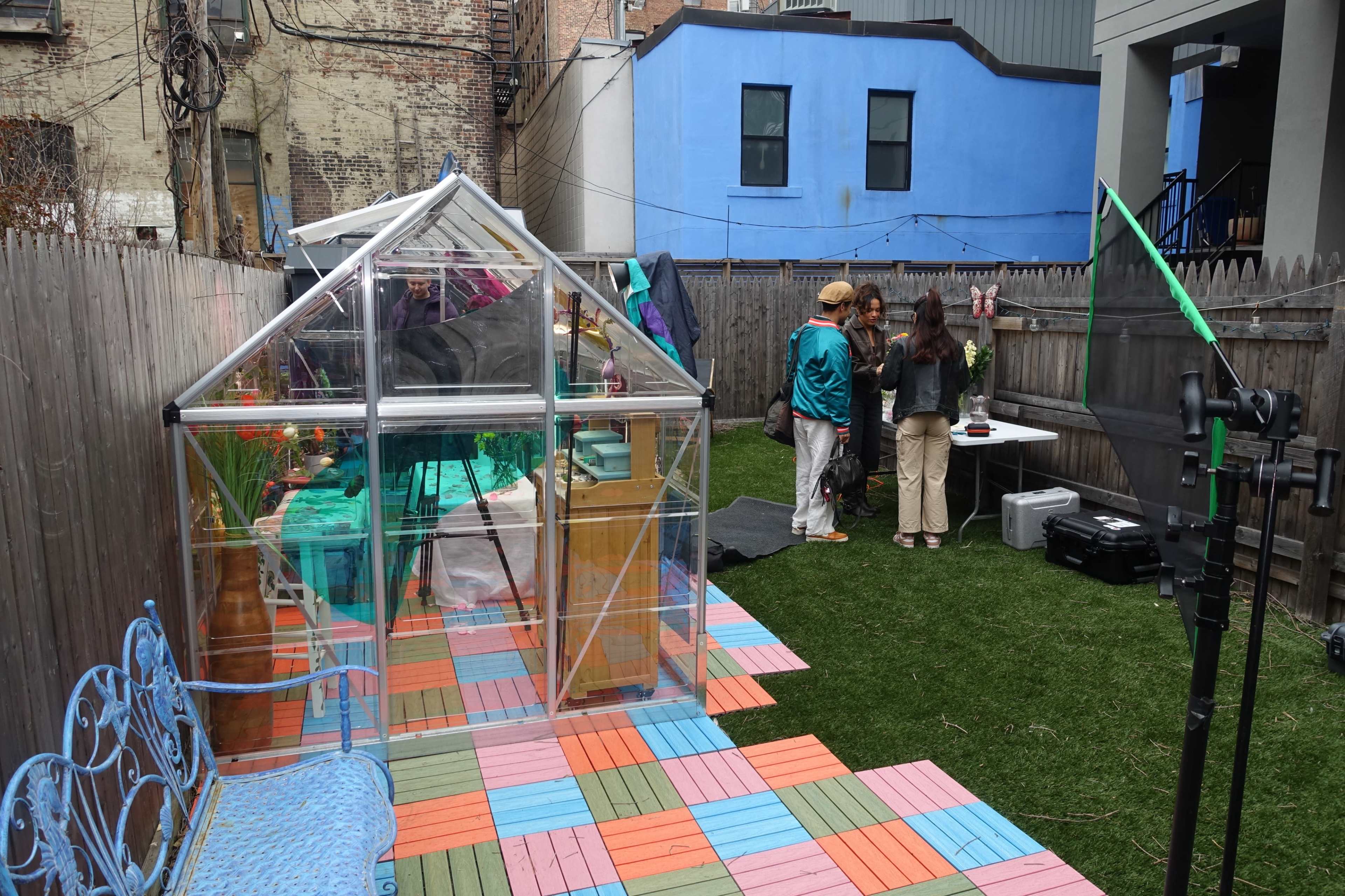 The image shows a backyard with a clear greenhouse, a colorful wooden patio, and a group of four people gathered around a table.