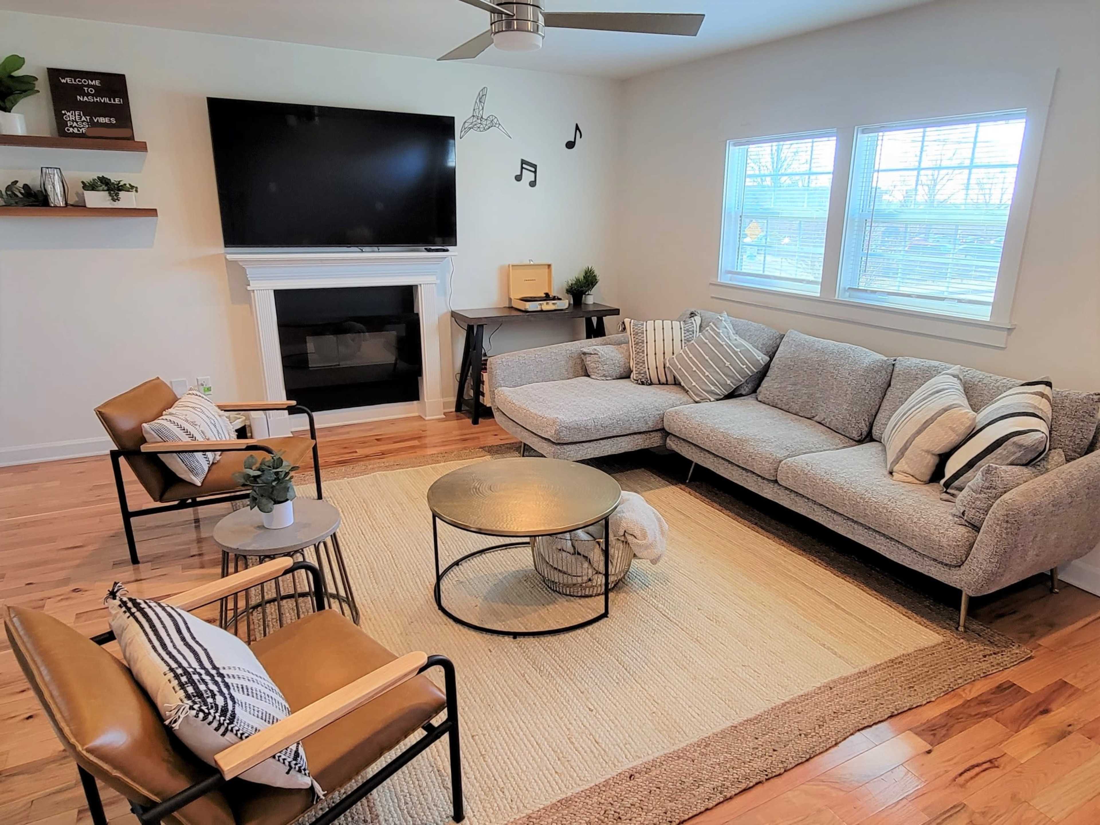 The living room features a gray sectional sofa, two brown armchairs, a round coffee table, and a fireplace, all arranged around a beige rug.
