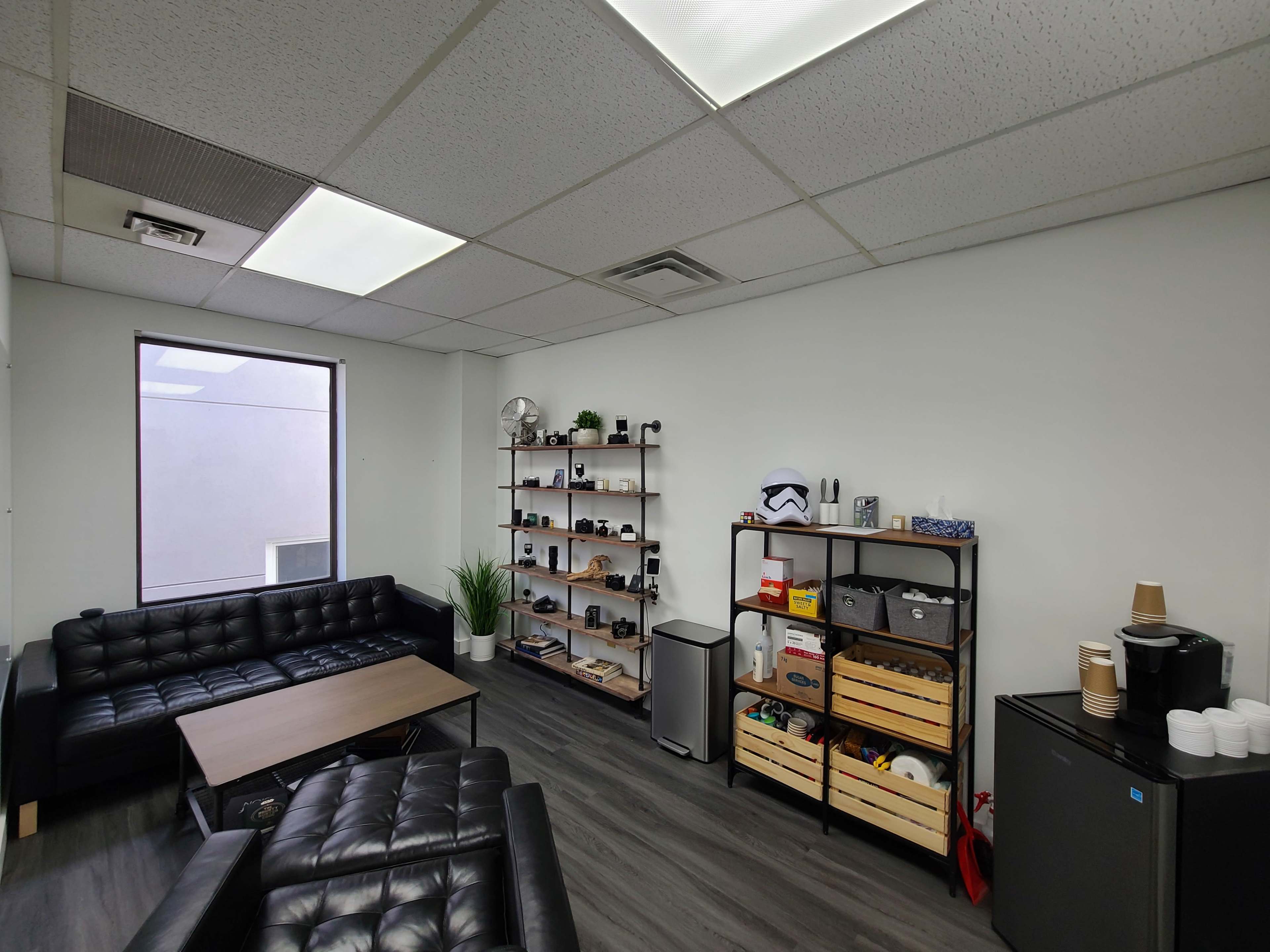 A modern office lounge with black leather sofas, a wooden coffee table, and shelves displaying various items, alongside a mini fridge and coffee station.