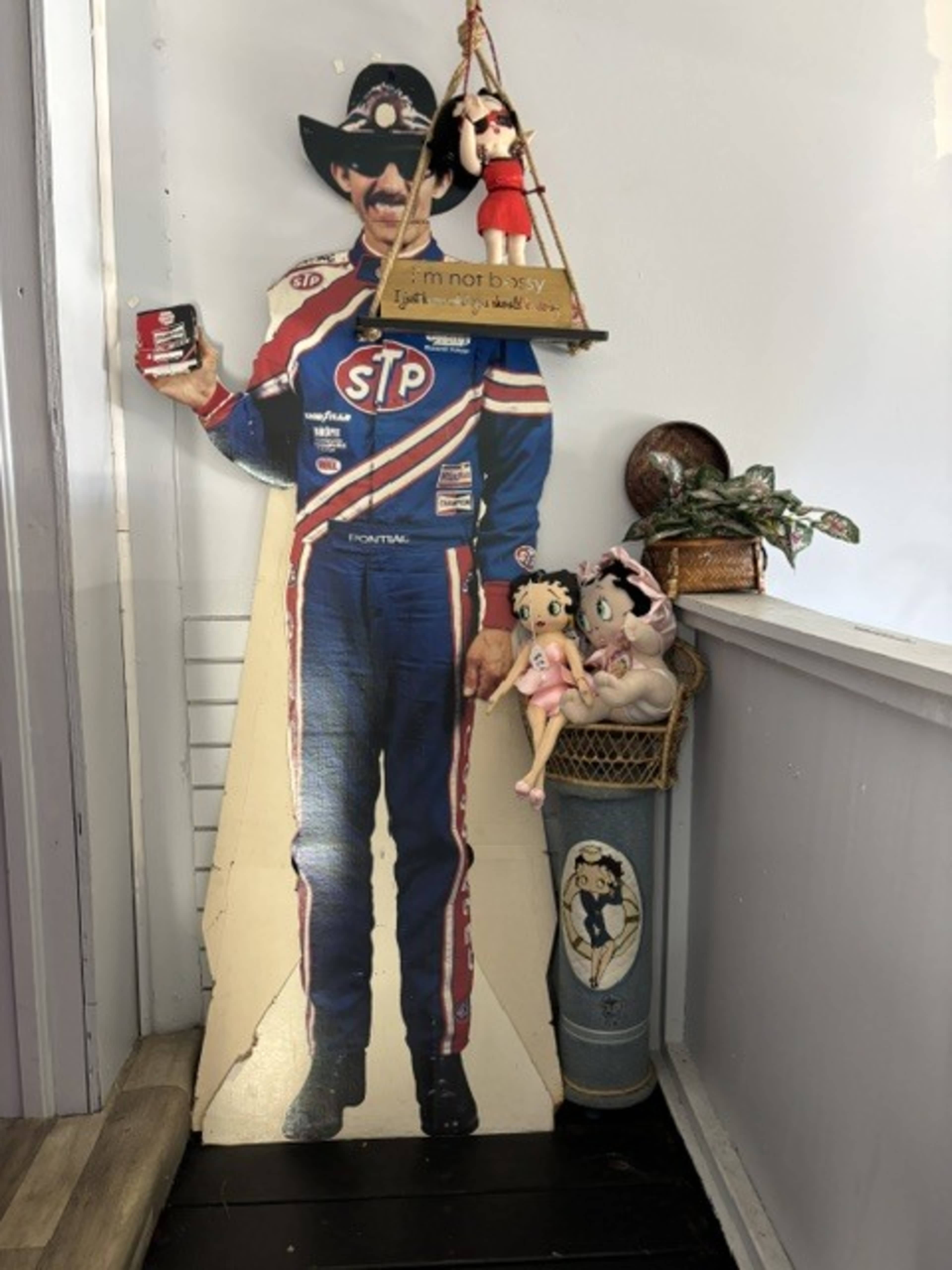 A life-sized cardboard cutout of a race car driver stands in a hallway alongside a basket of dolls and a decorative shelf.