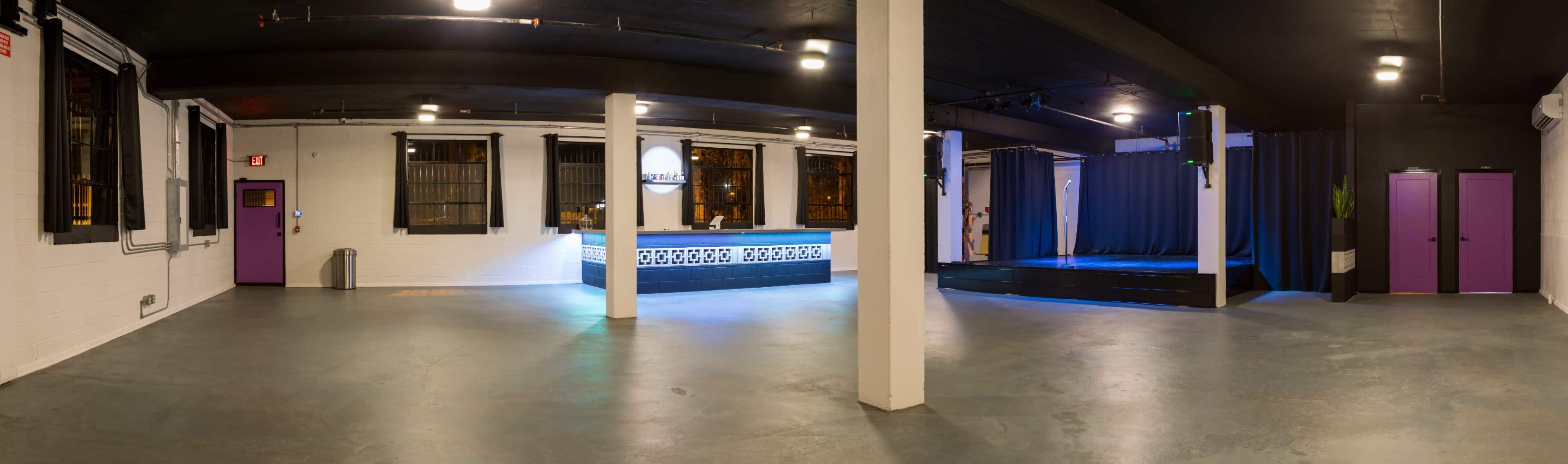 An interior of a spacious venue with a bar area on the left and a stage with curtains on the right, illuminated in soft lighting.