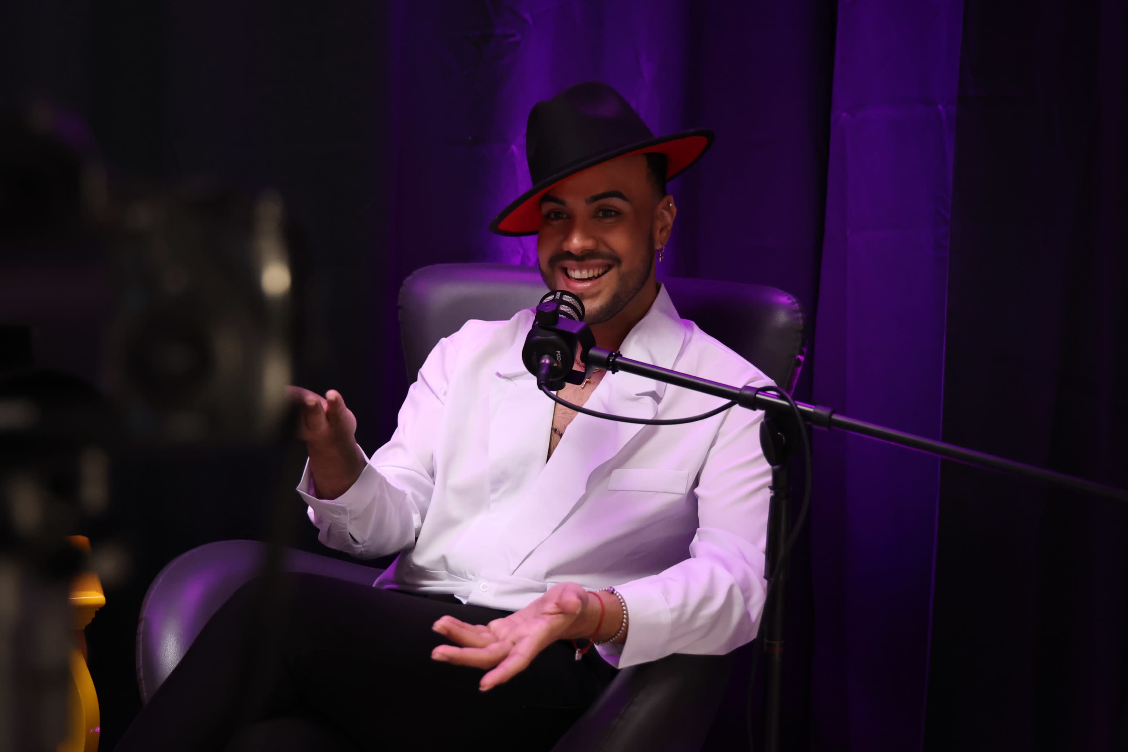 A person wearing a white shirt and a black hat sits in a chair, smiling and gesturing while speaking into a microphone against a purple backdrop.