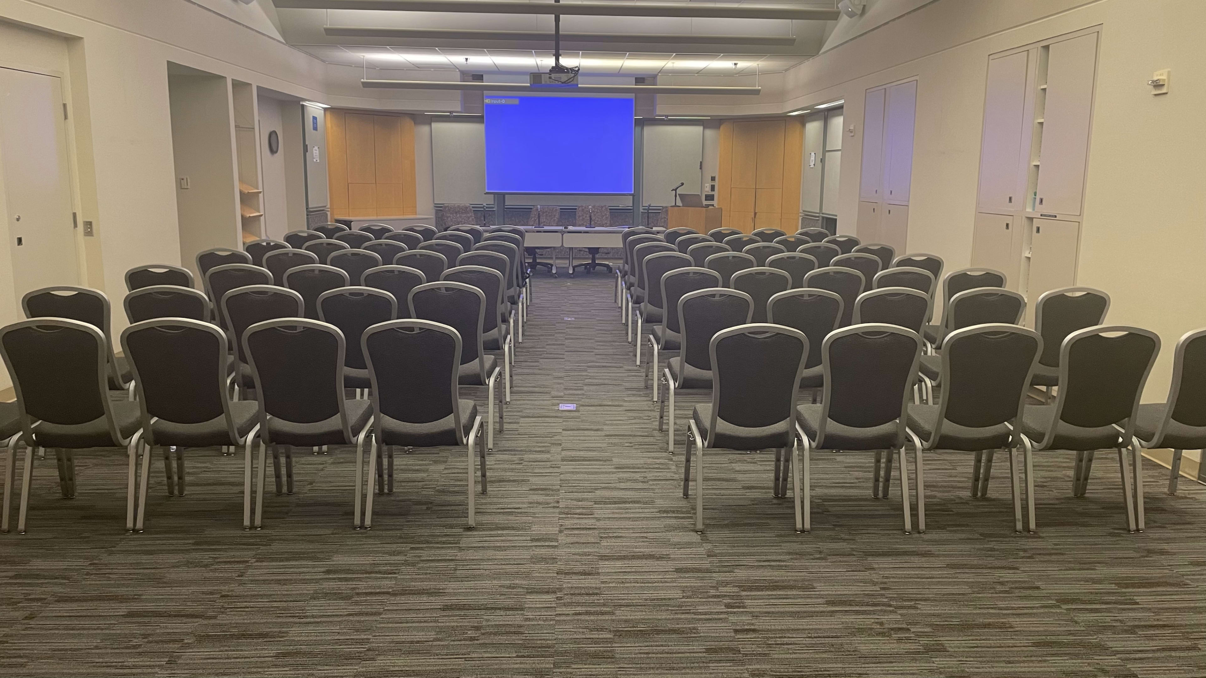 A conference room with rows of black chairs facing a large blank screen.