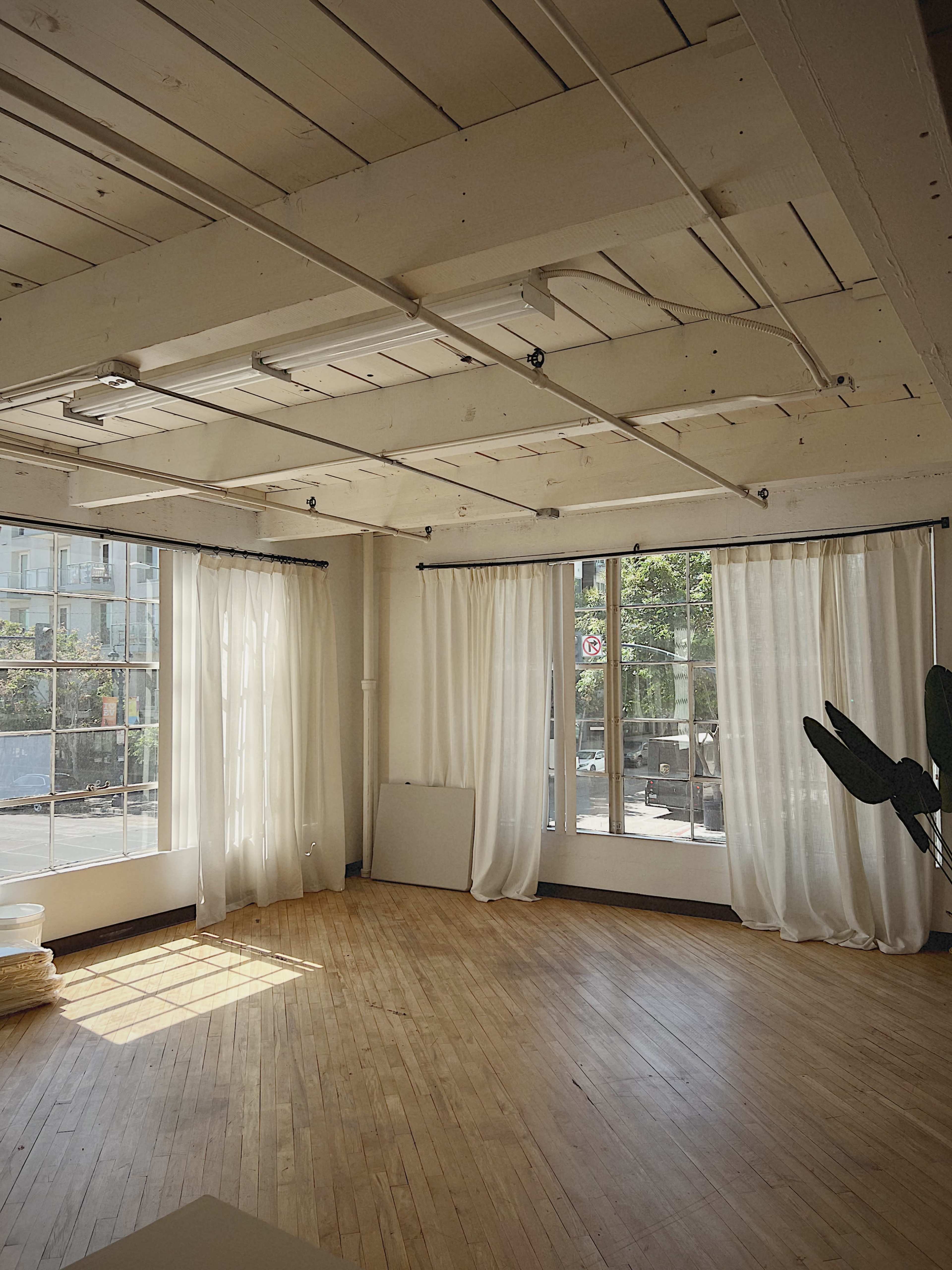 The image shows a spacious room with wooden floors, large windows draped with sheer white curtains, and natural light streaming in.