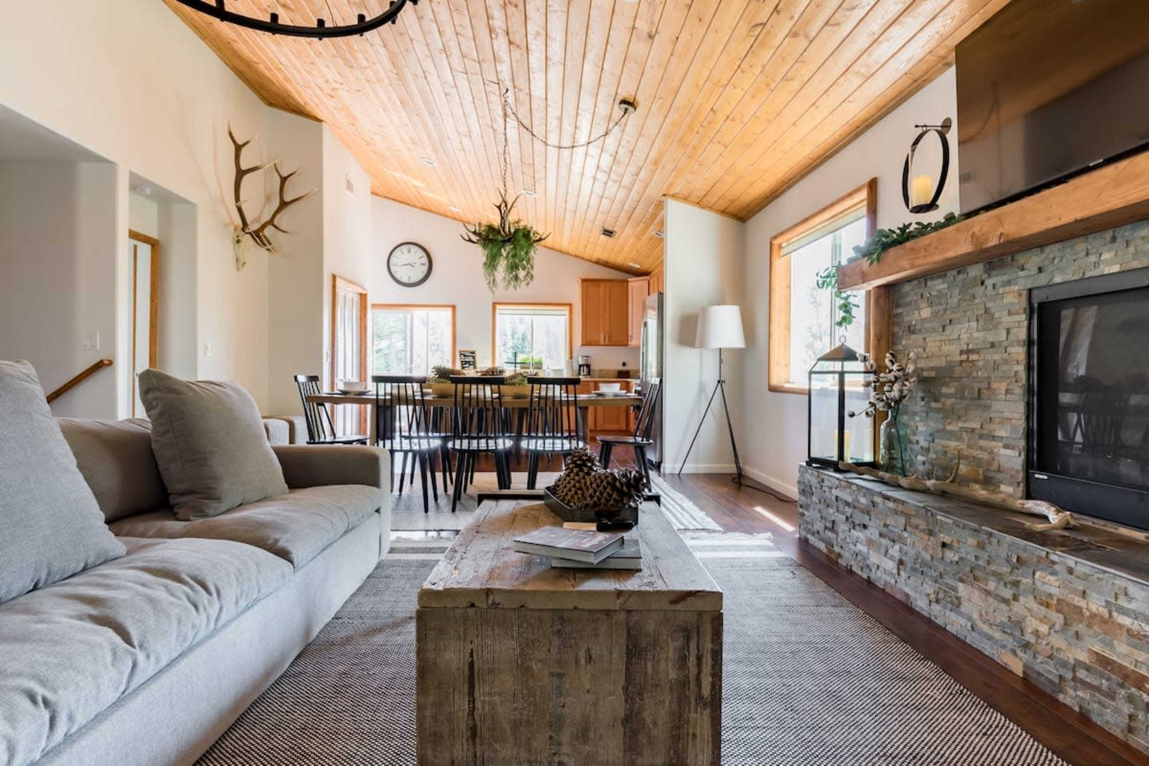 The image shows a modern living room with a wooden ceiling, a stone fireplace, a sofa, a coffee table, and a dining area visible in the background.