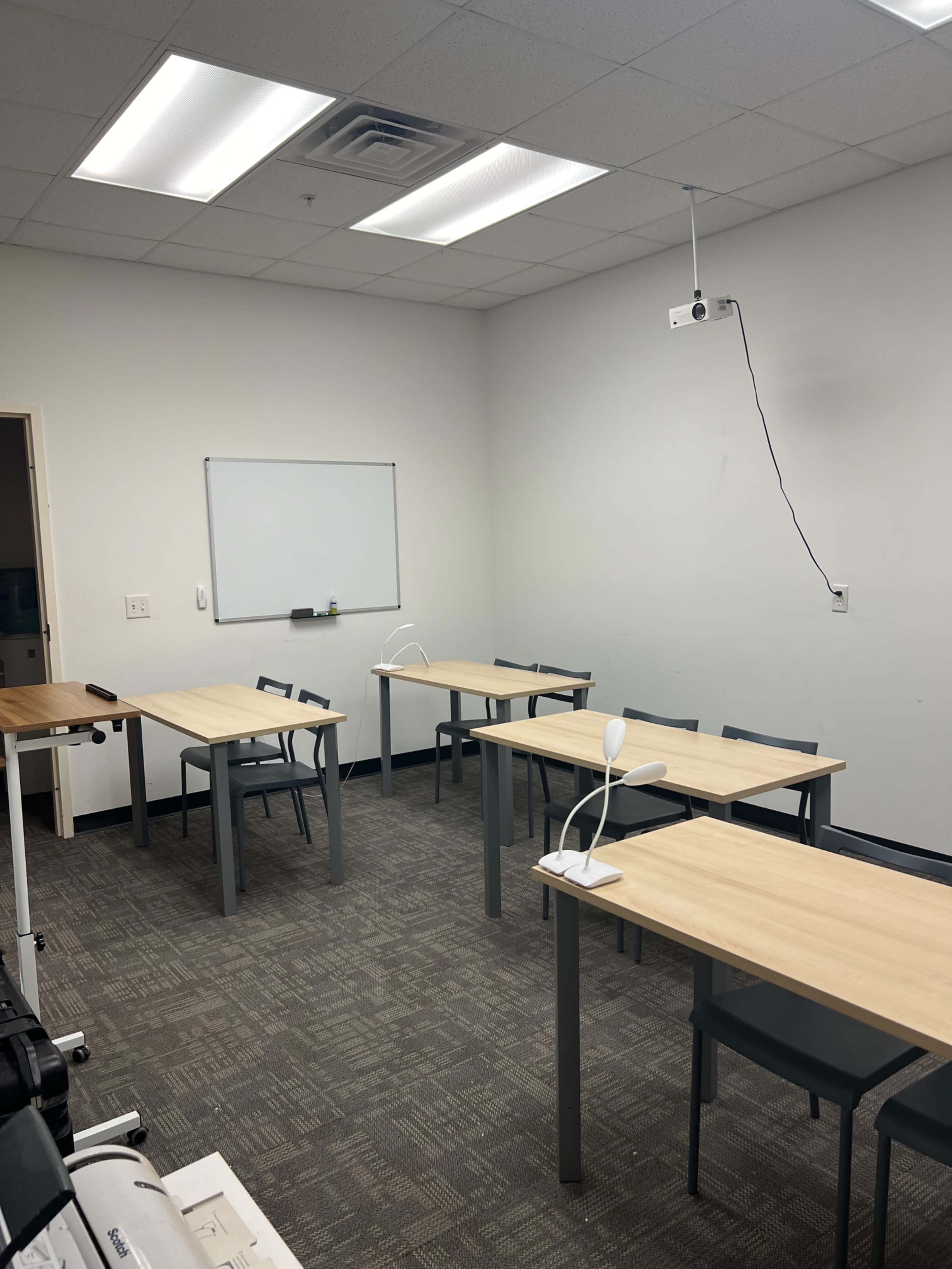 A classroom with five tables arranged in a U-shape, a whiteboard on the wall, and a projector hanging from the ceiling.