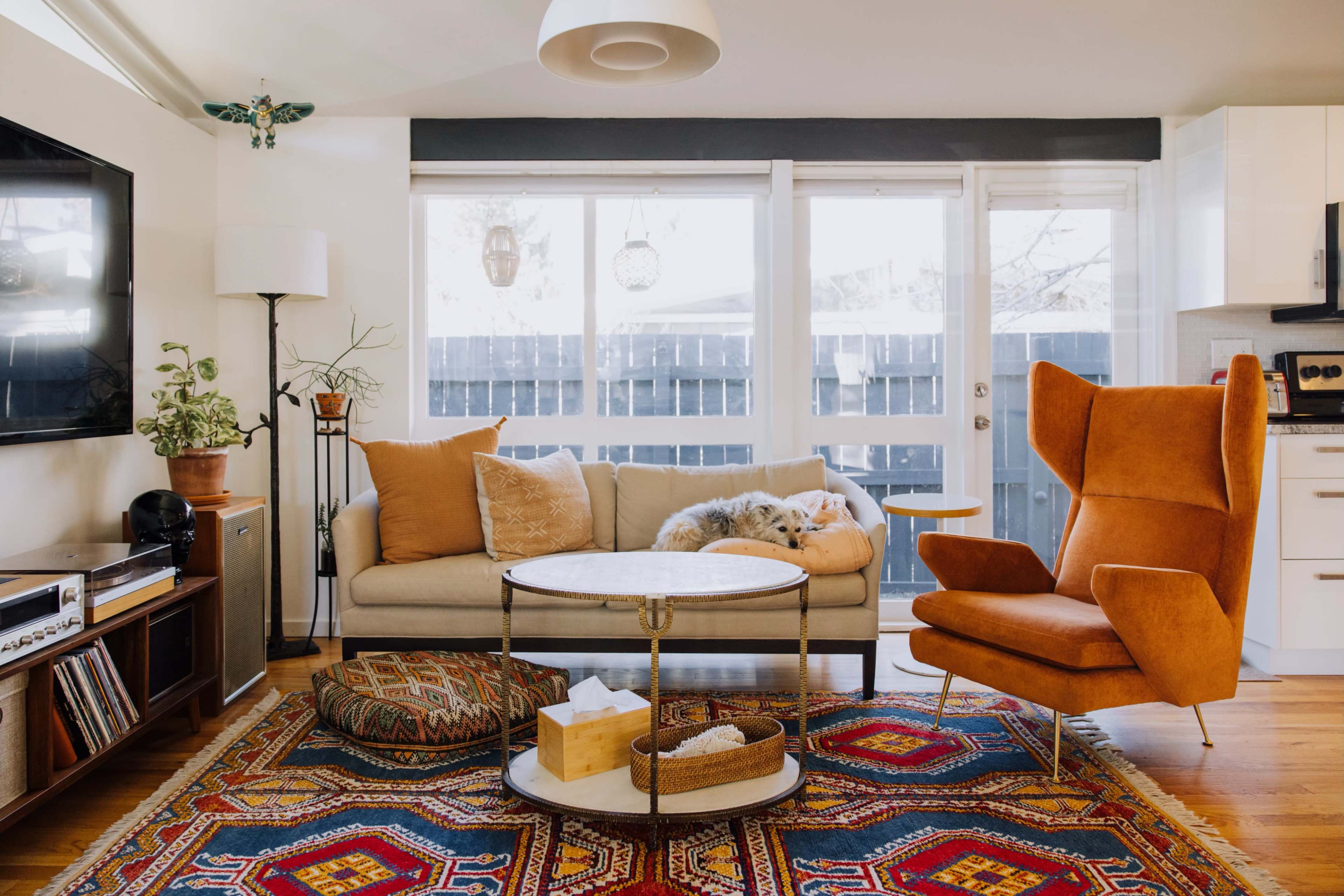 A cozy living room features a beige sofa, an orange accent chair, and a round coffee table, all set against a backdrop of large windows and a vibrant area rug.