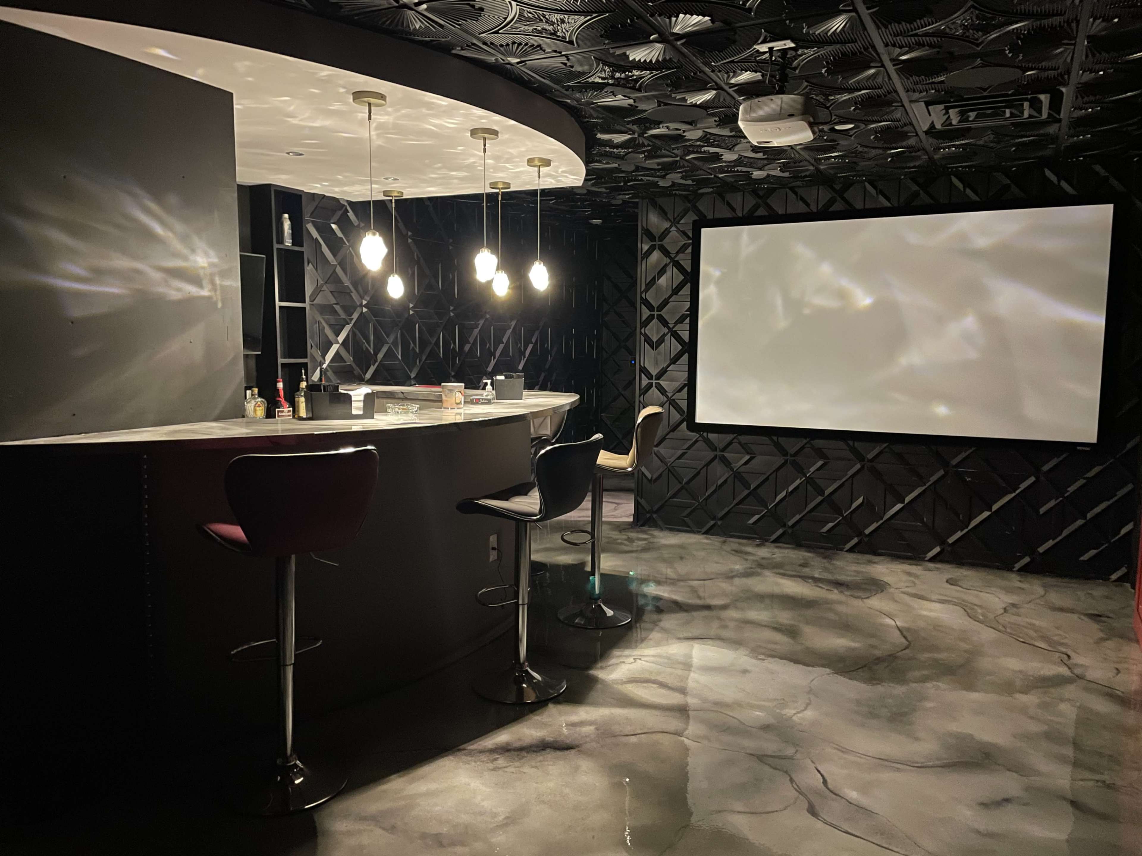 A stylish room with a bar area featuring modern stools and a large blank screen on one wall, illuminated by pendant lights.
