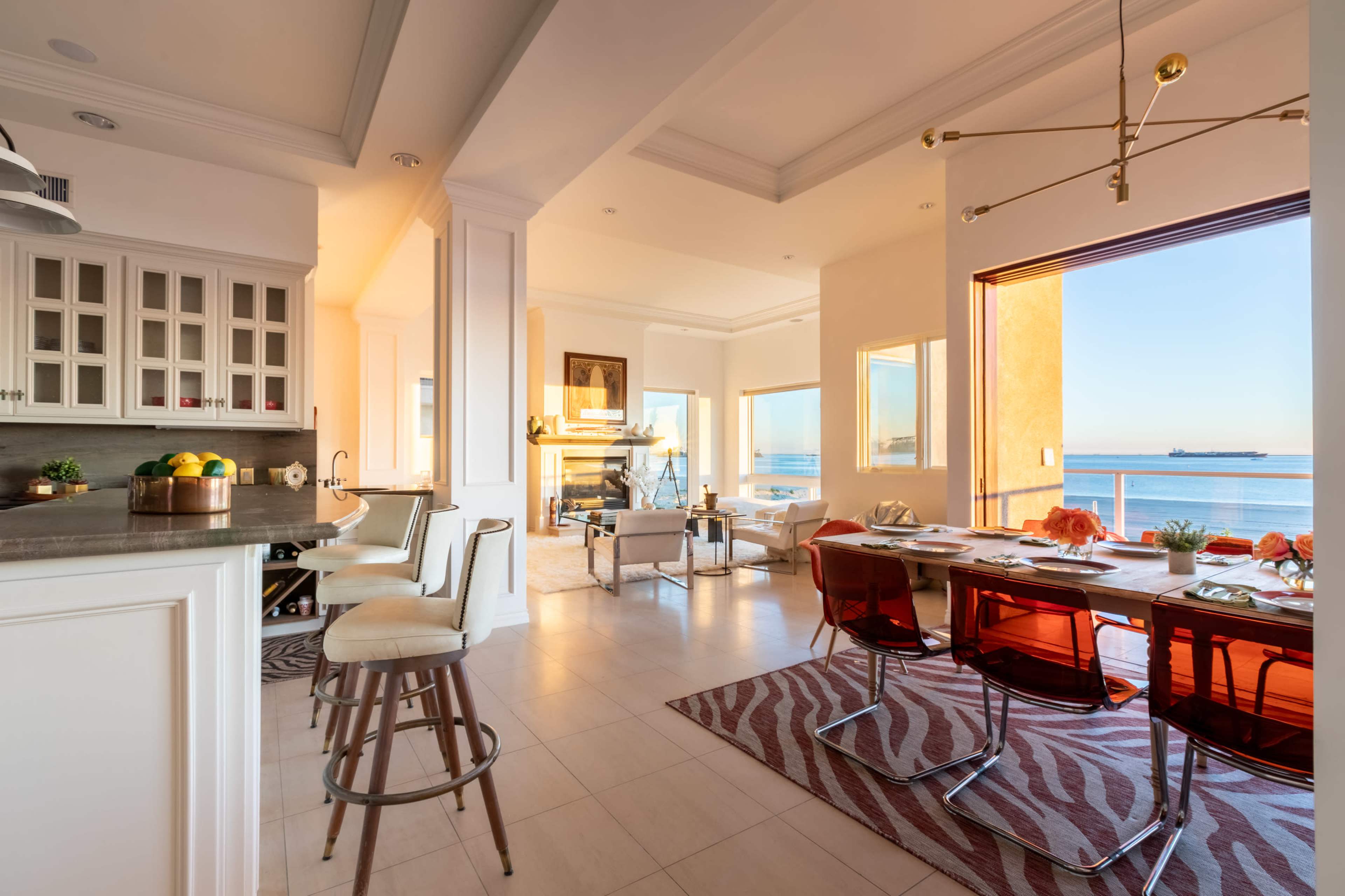 The image shows a modern, bright open-concept kitchen and dining area with a view of the water and large windows.