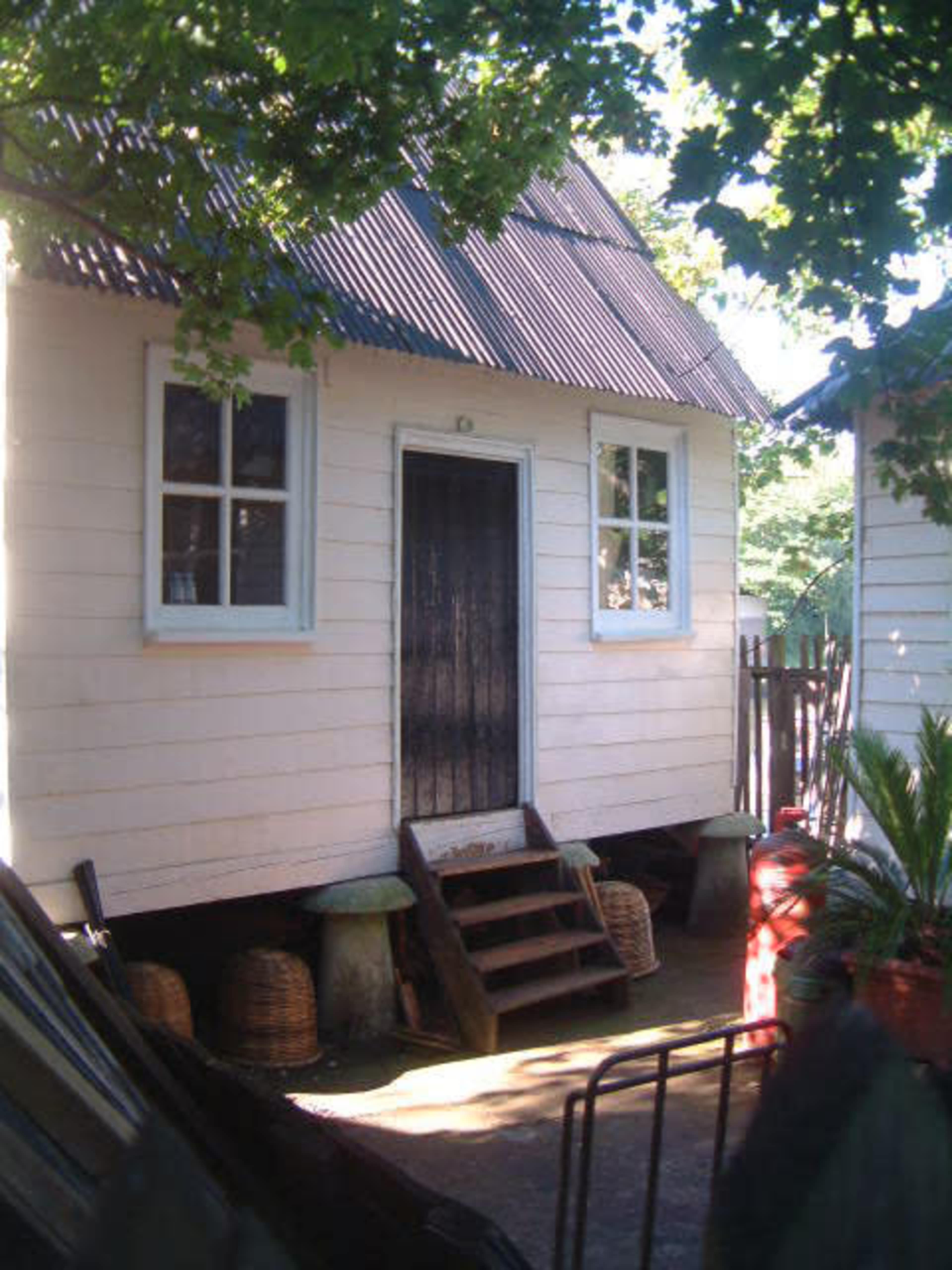 A small white wooden house with a dark door and a sloped metal roof, surrounded by trees and outdoor furnishings.