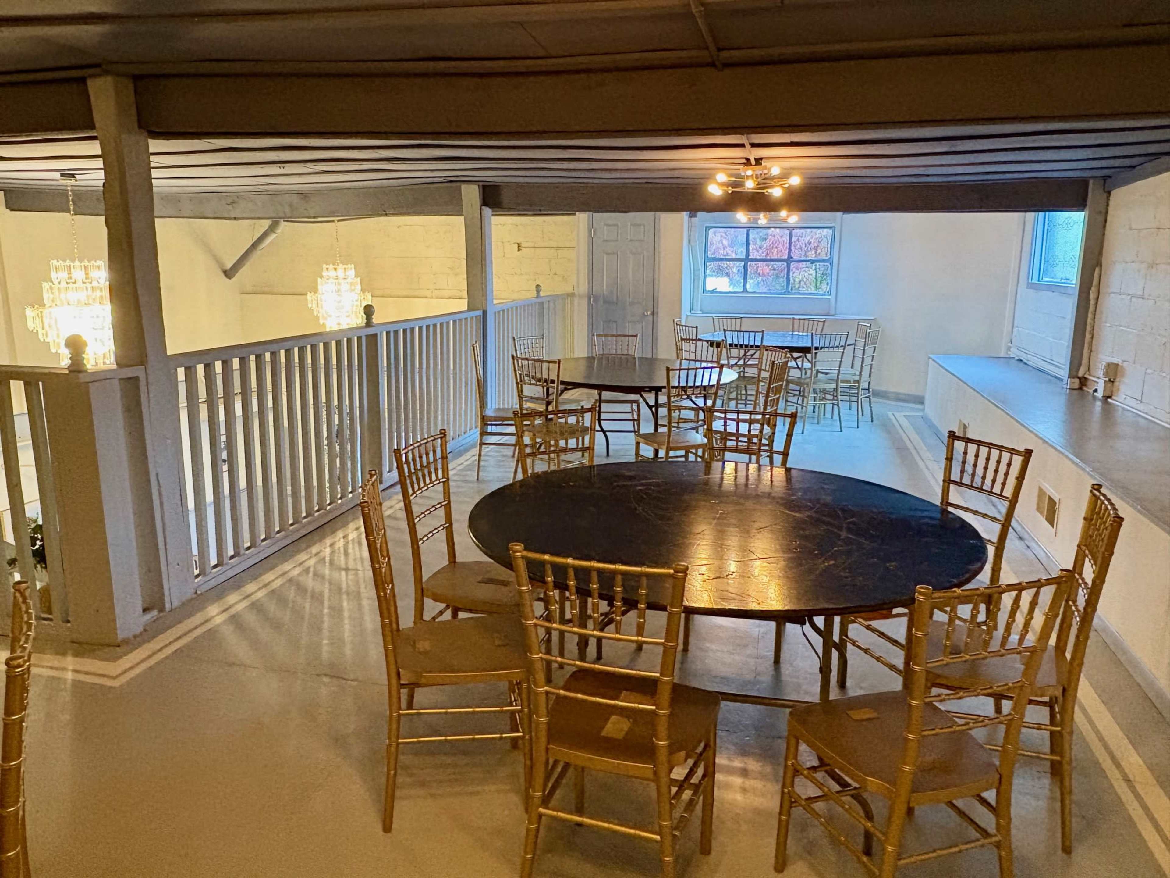 The image shows a spacious room with several round and rectangular tables surrounded by gold chiavari chairs, set in a well-lit area with white walls and wood beams.