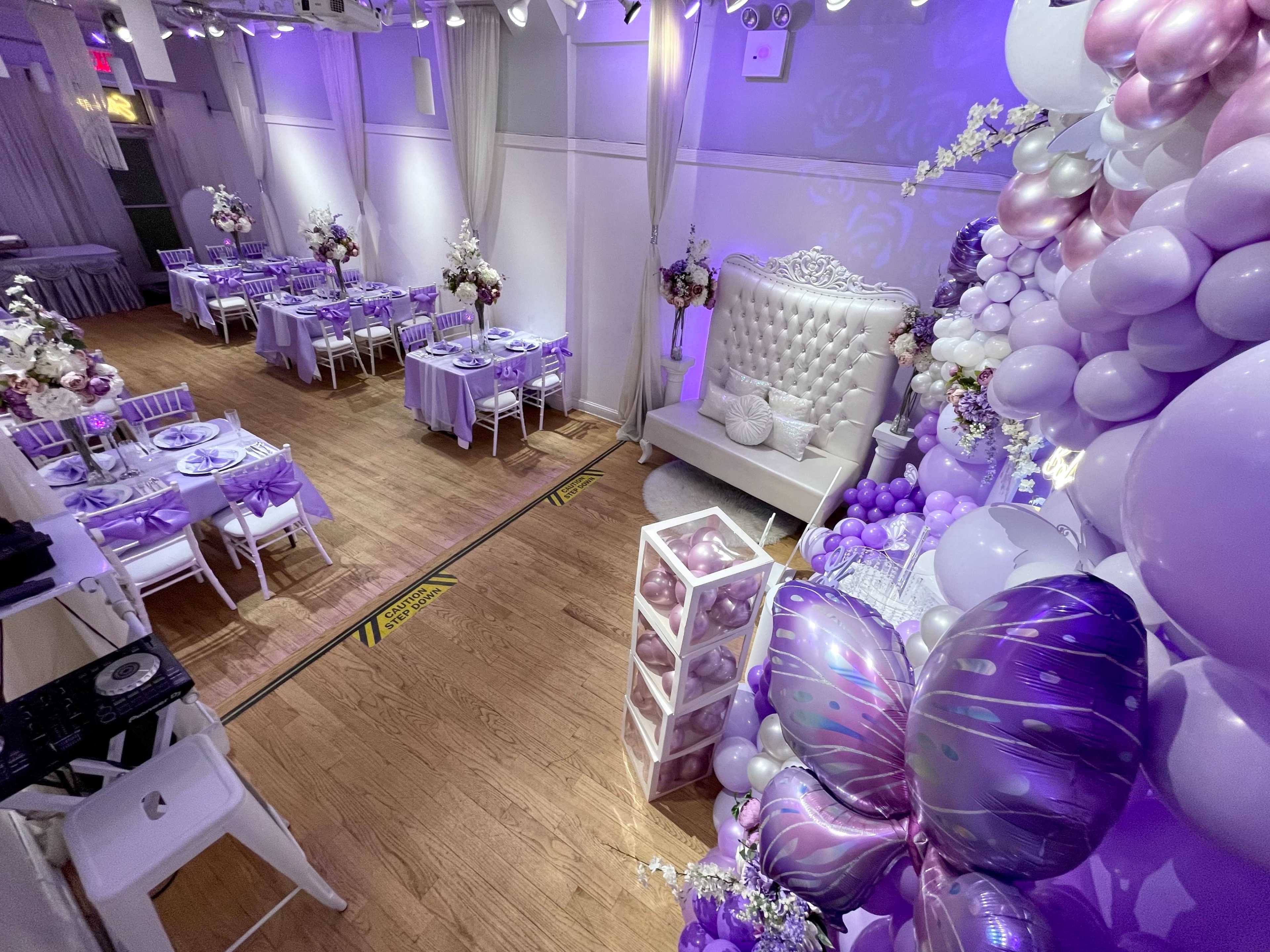The image showcases a decorated event space with tables set up for a celebration, featuring purple and pink balloons, floral arrangements, and a plush seating area.