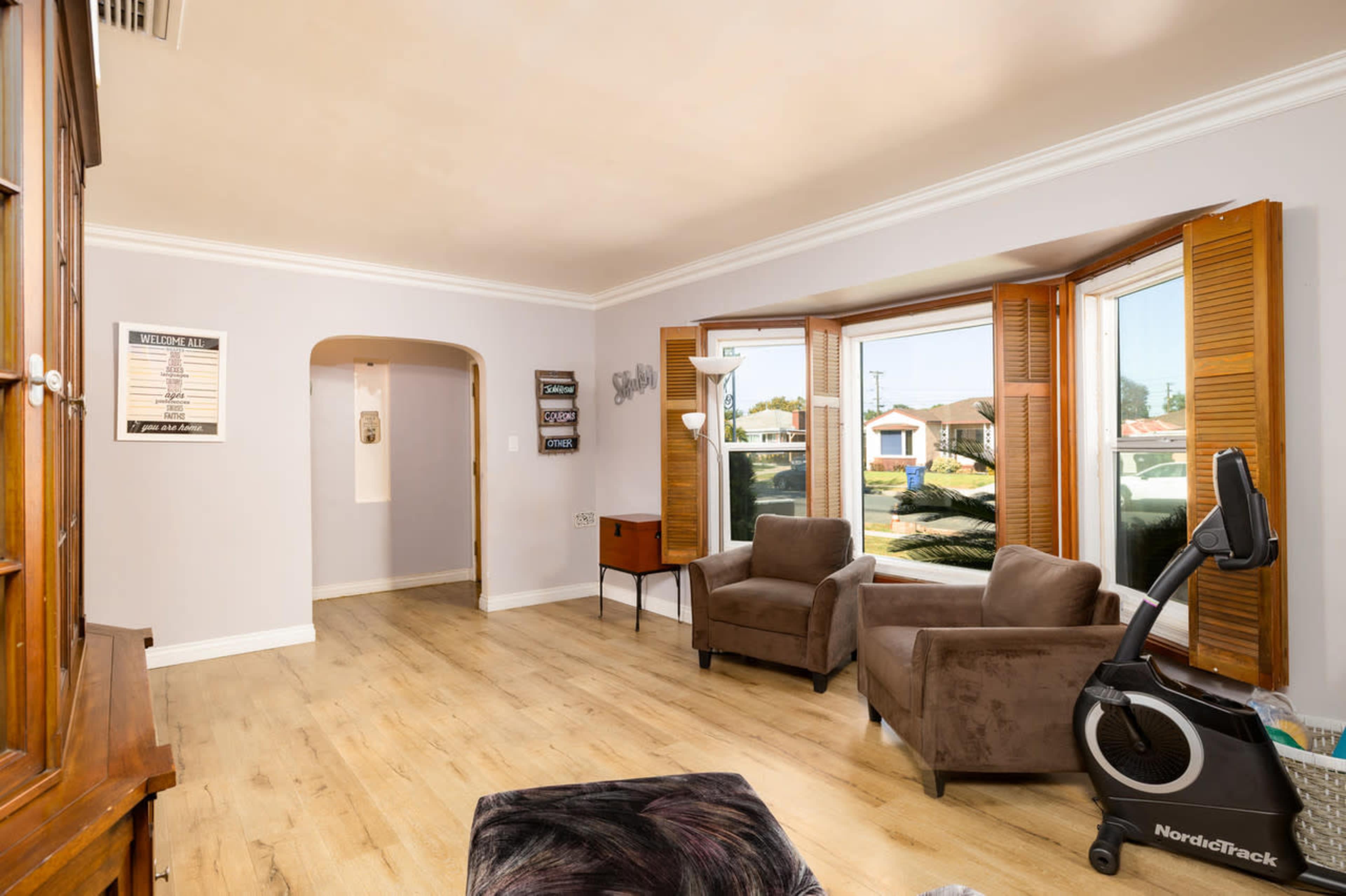 The image shows a light-colored living room with two brown armchairs, a wooden cabinet, and a large window with wooden shutters.