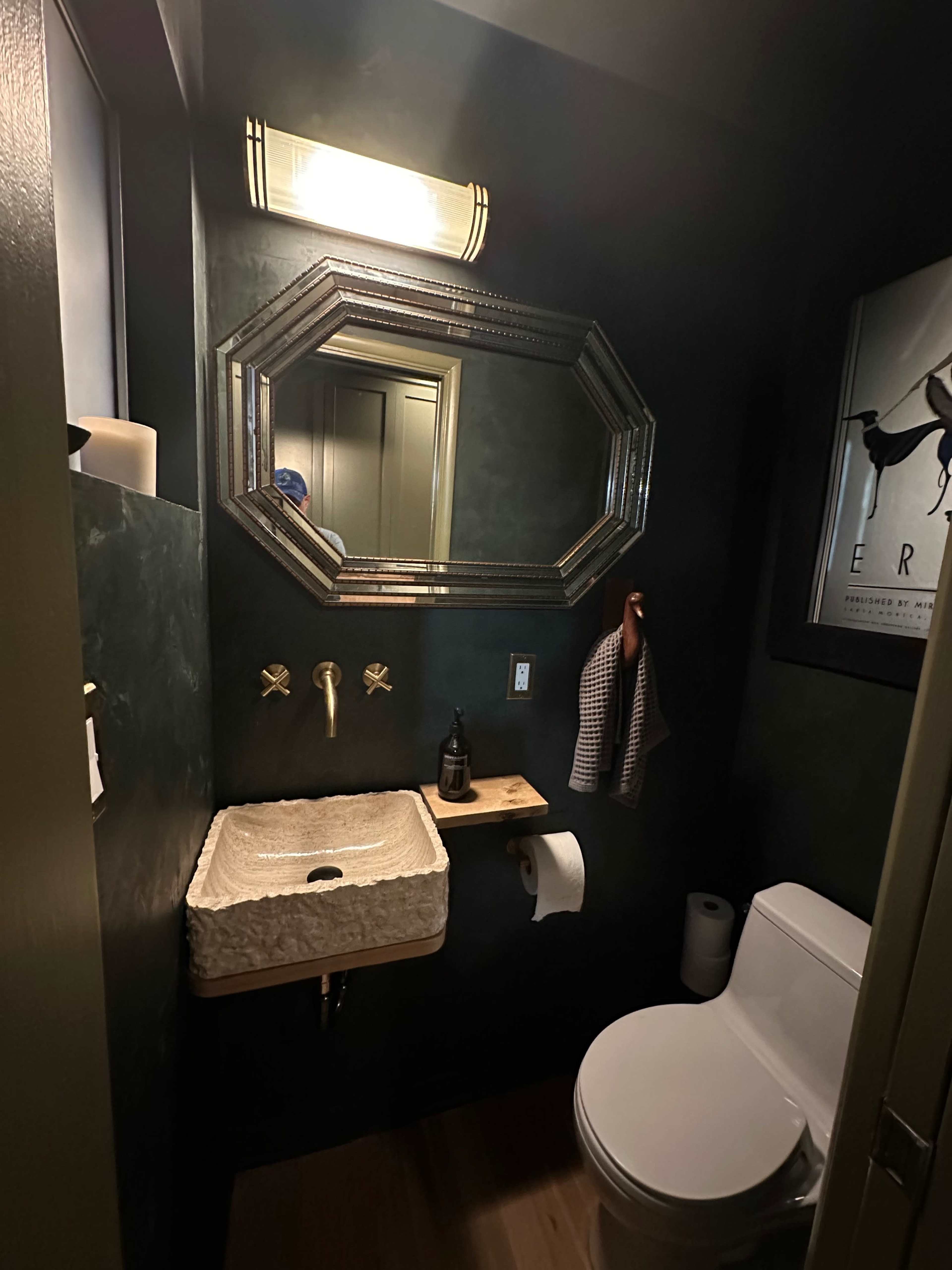 The image shows a small bathroom with a dark green wall, a wall-mounted sink with a wooden shelf, a modern toilet, and decorative elements including a mirror and artwork.