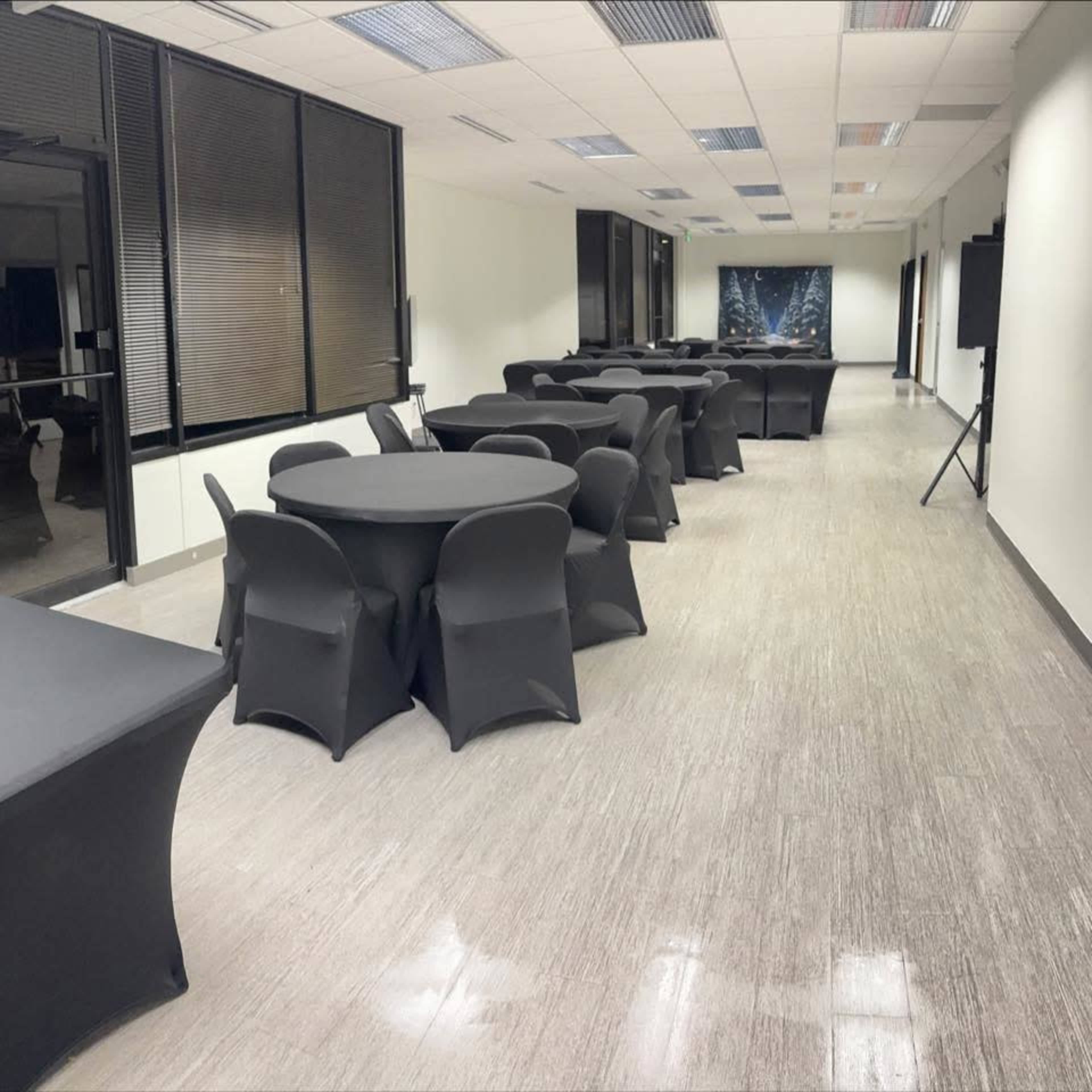 A row of black-covered round tables is arranged in a brightly lit, empty conference room with large windows.