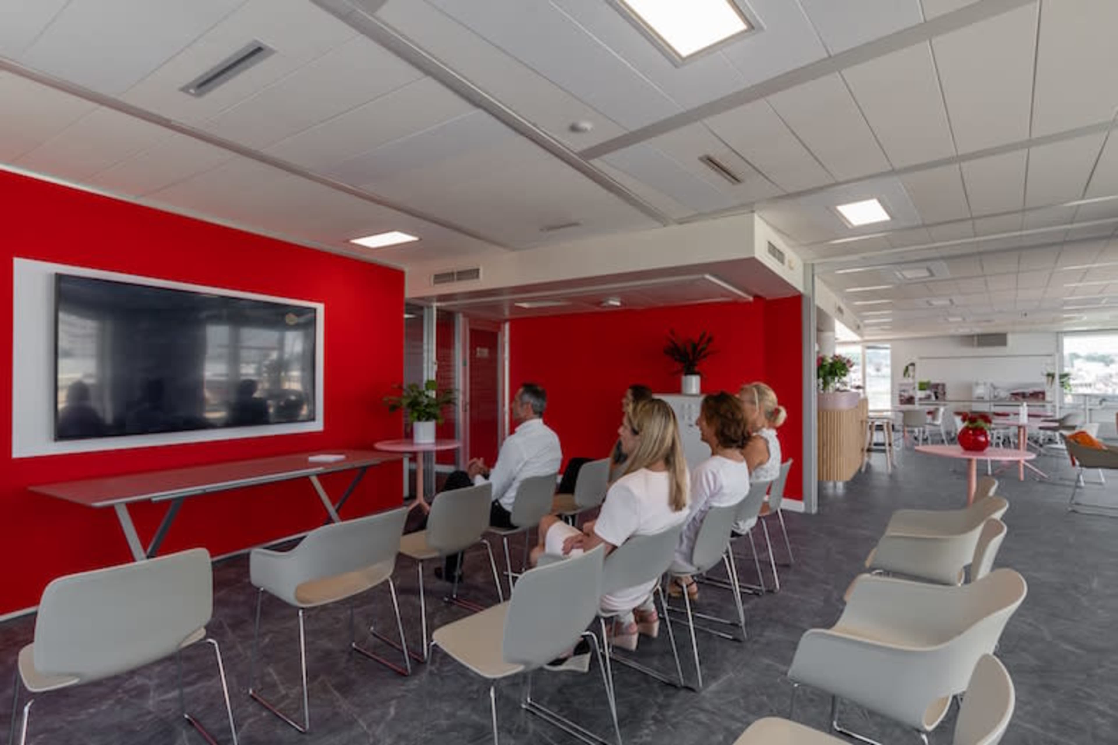 A group of people sits in a modern office space facing a large television mounted on a red wall.