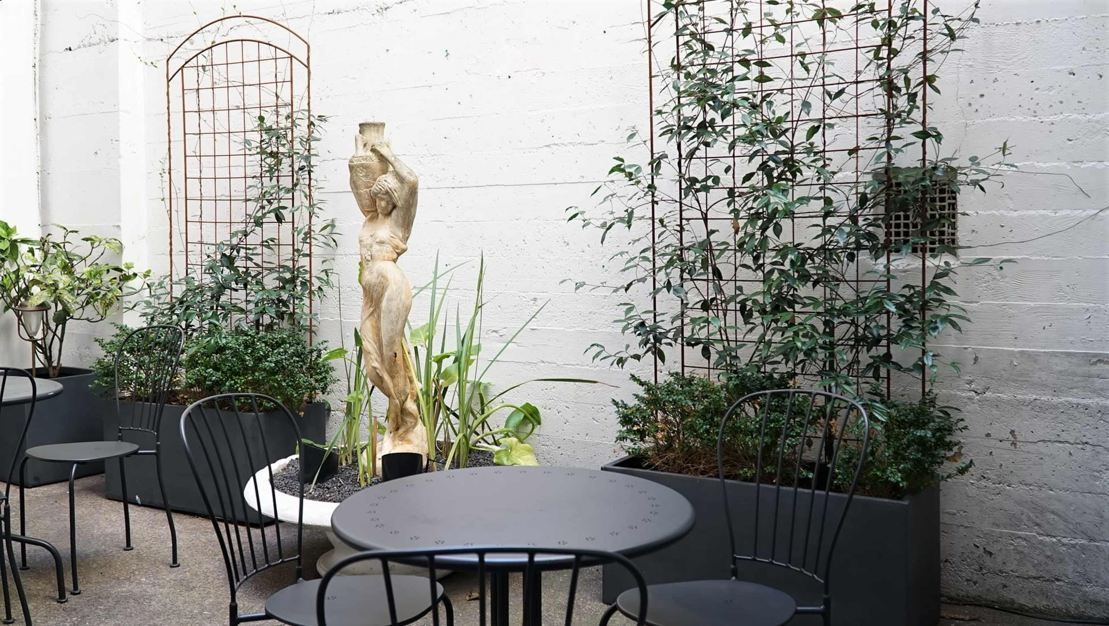 A modern outdoor seating area with black tables and chairs, flanked by tall planters with greenery and a wooden sculpture of a figure.