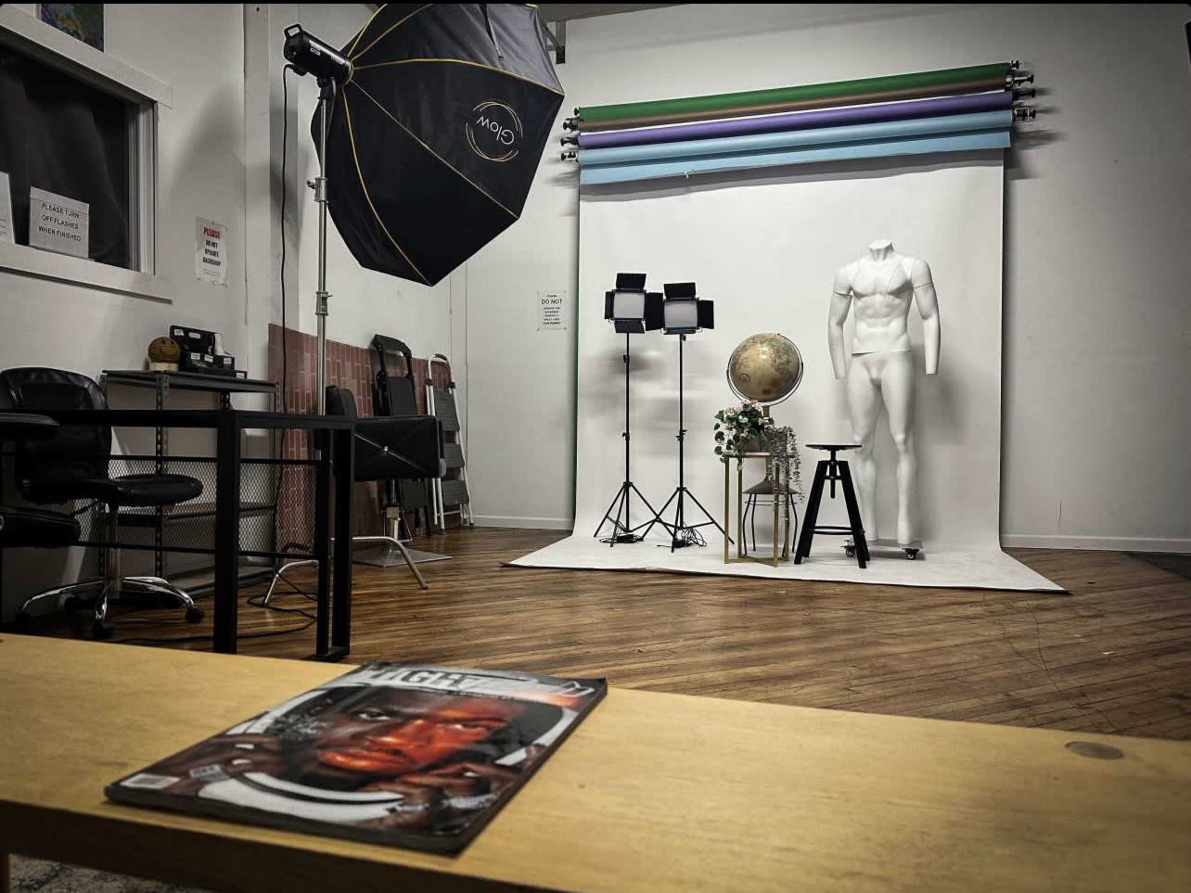 The image shows a photography studio with a backdrop of colored paper, a mannequin, a globe, and various lighting equipment, while a magazine rests on a table in the foreground.