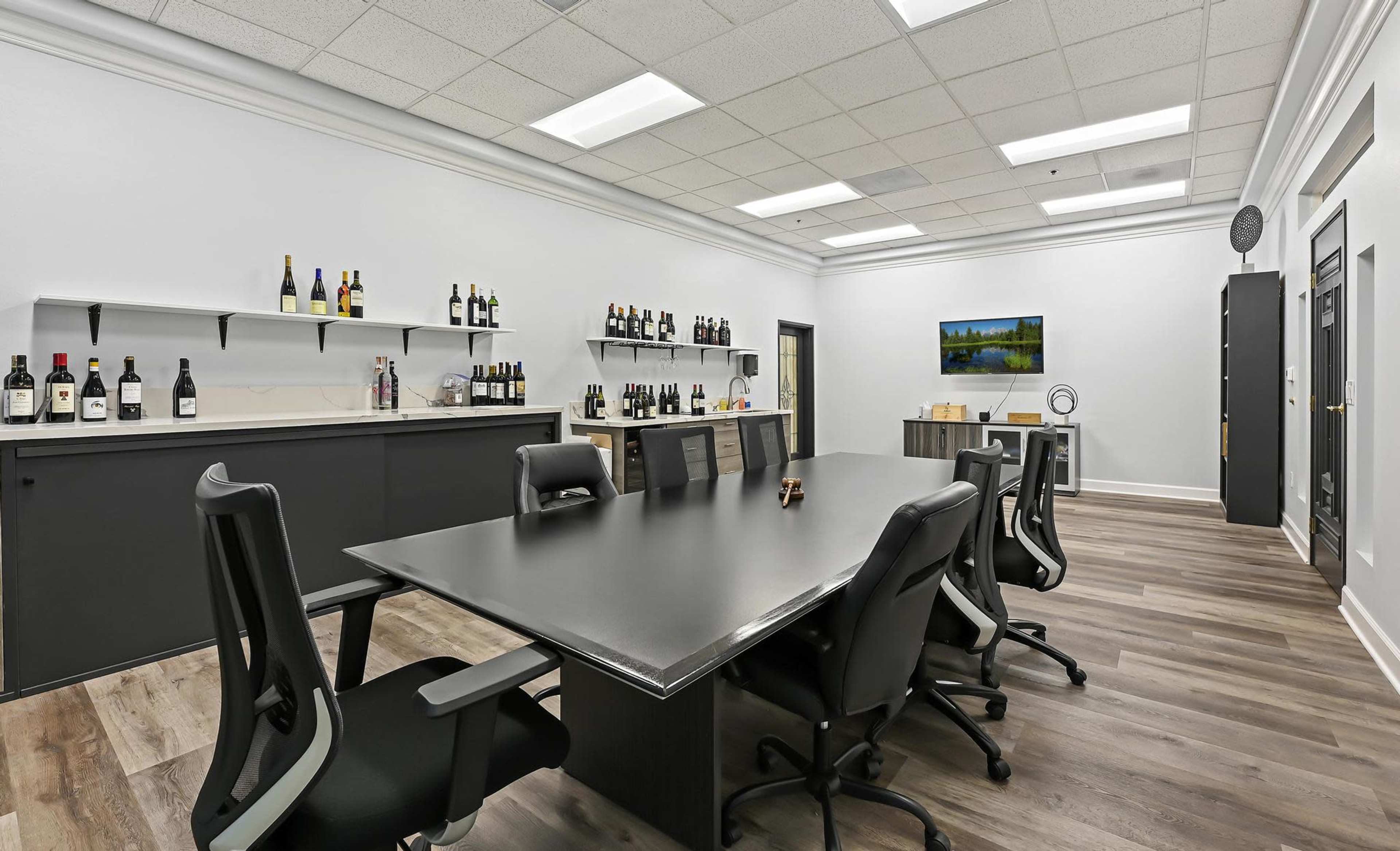 A modern conference room with a large black table, black ergonomic chairs, and shelves lined with various bottles, complemented by a television on the wall.