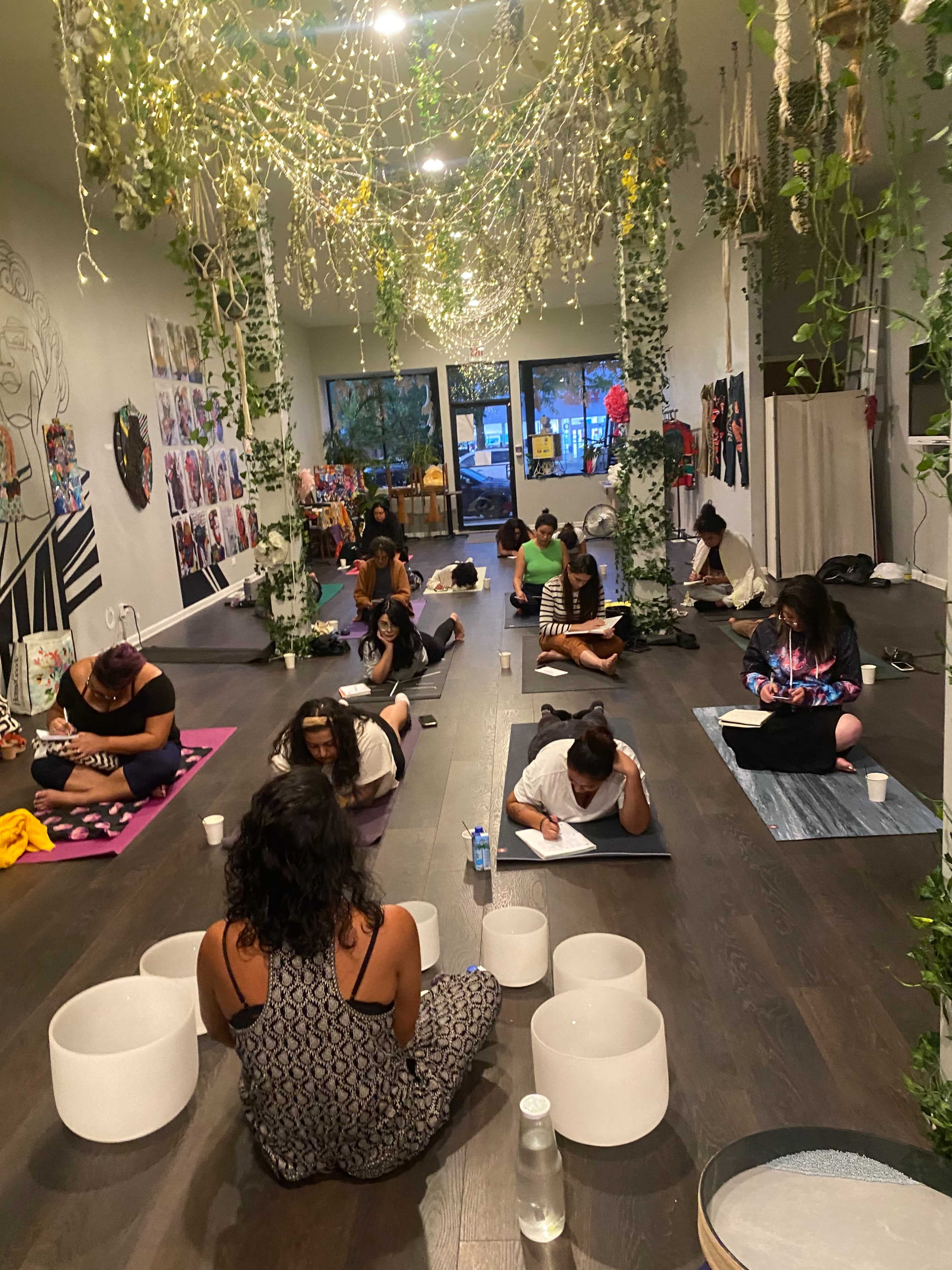 A group of people sit on yoga mats in a dimly lit studio adorned with plants and soft lighting, engaged in a drawing or meditation activity.