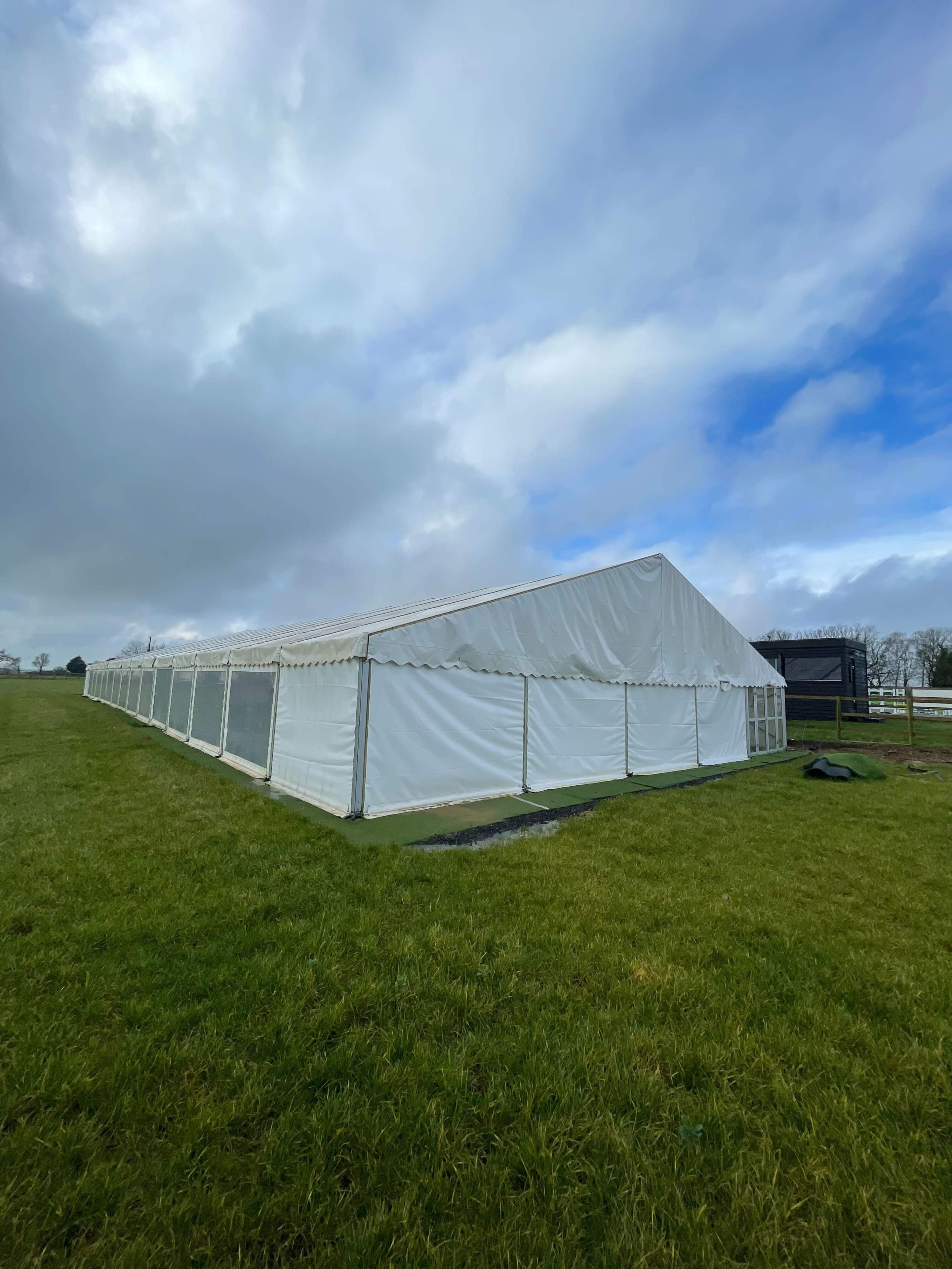 A large white tent with side panels is set up on a grassy field under a cloudy sky.