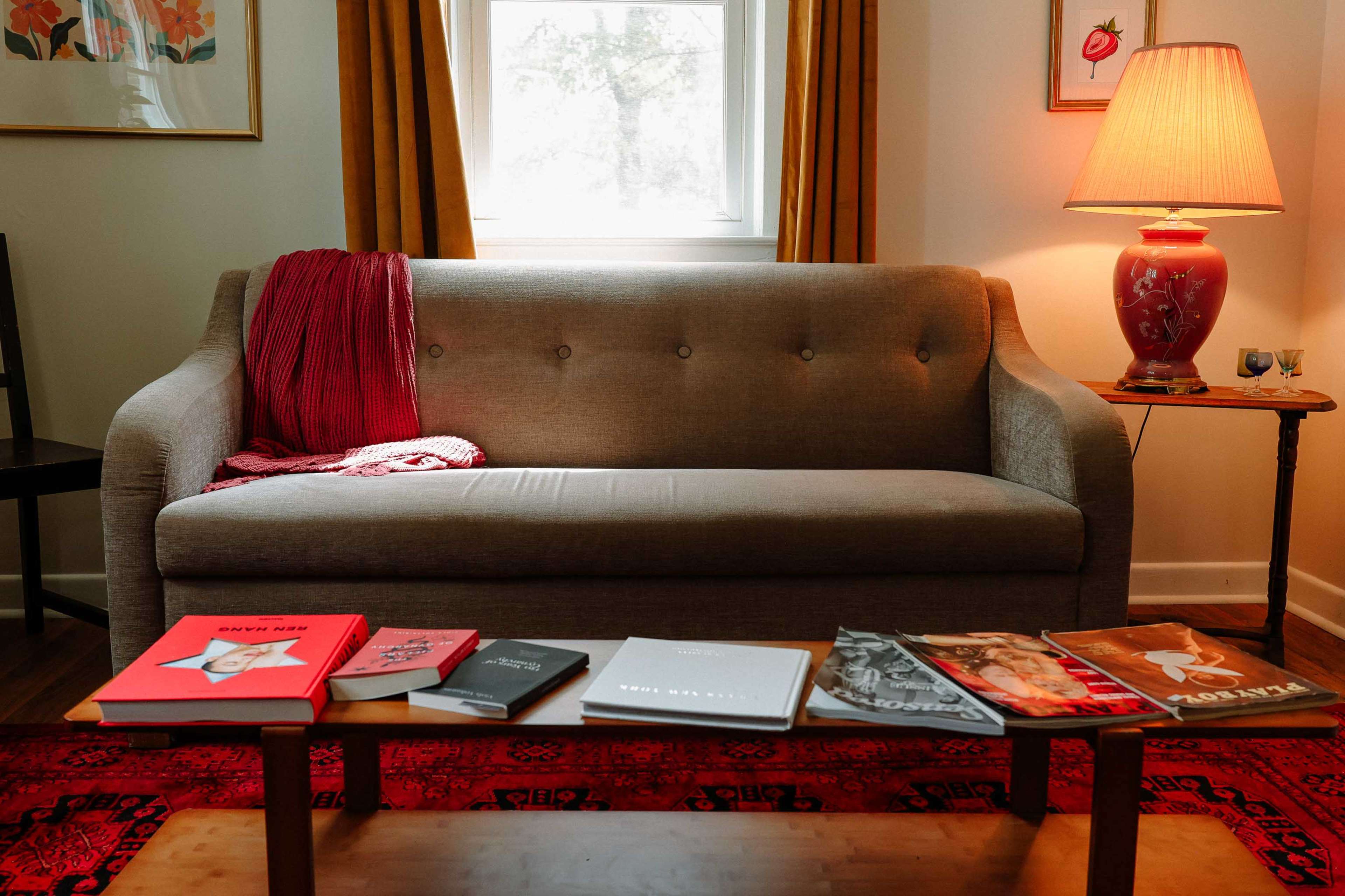 A gray couch with a red throw rests against a wall with a window, while a lamp illuminates a coffee table featuring several books and a patterned rug underneath.