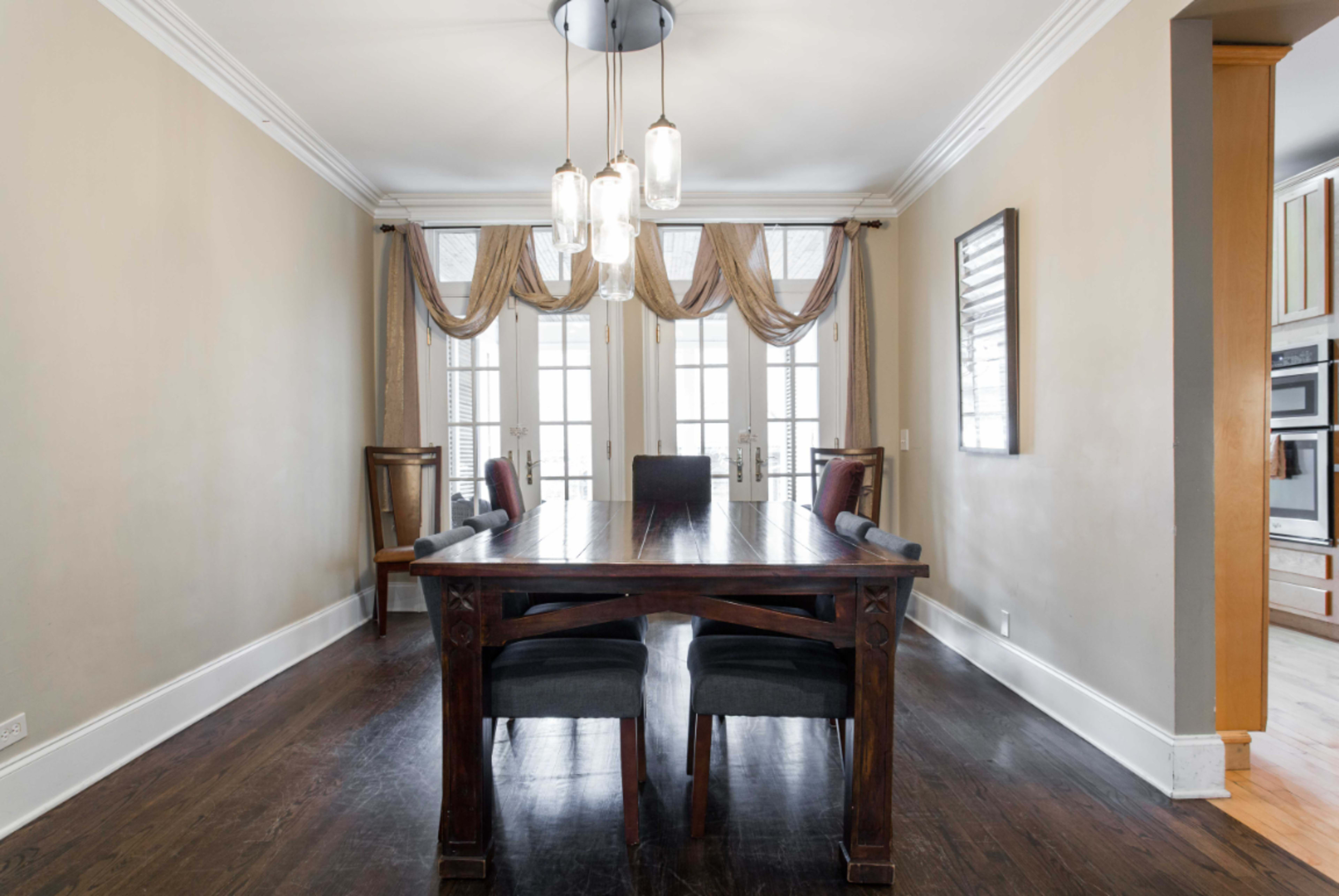 A dining room features a long wooden table with dark chairs, illuminated by a trio of pendant lights above, and flanked by large windows with draped curtains.