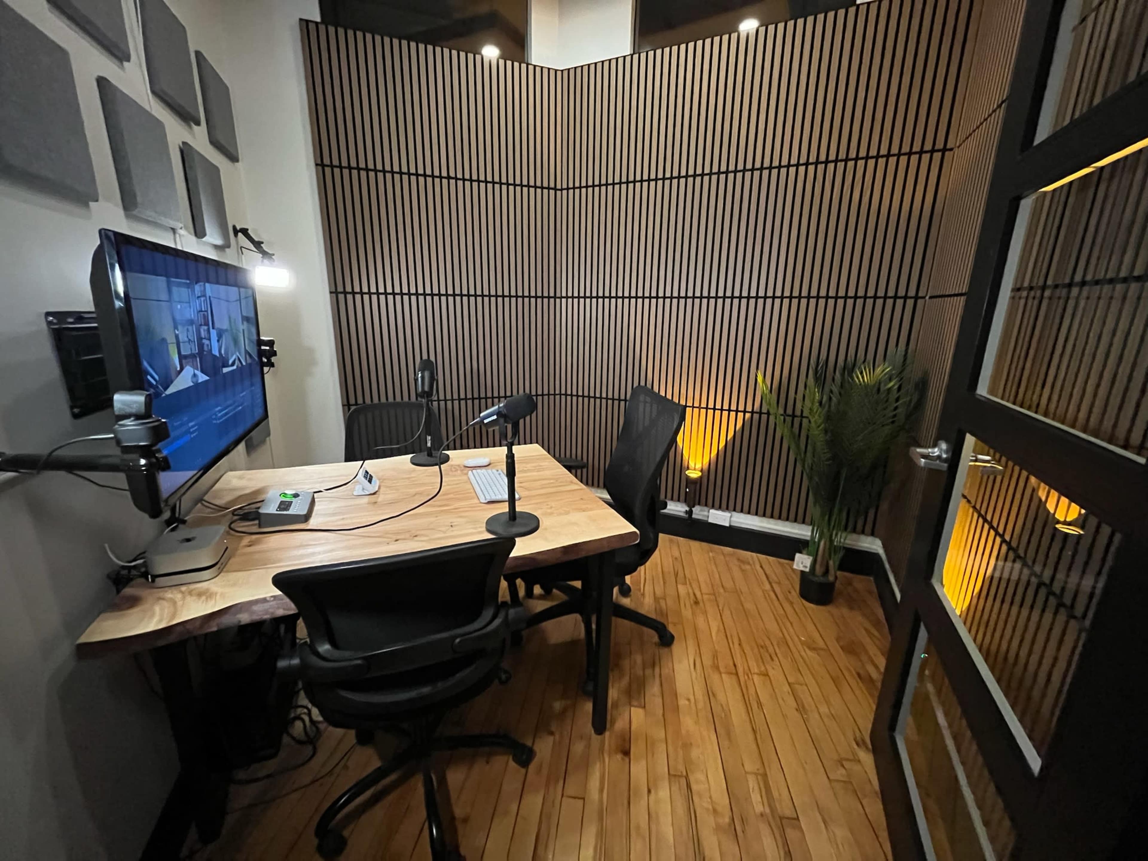 The image shows a small recording studio featuring a wooden table with microphones, a monitor displaying content, black chairs, and wooden paneling on the walls.