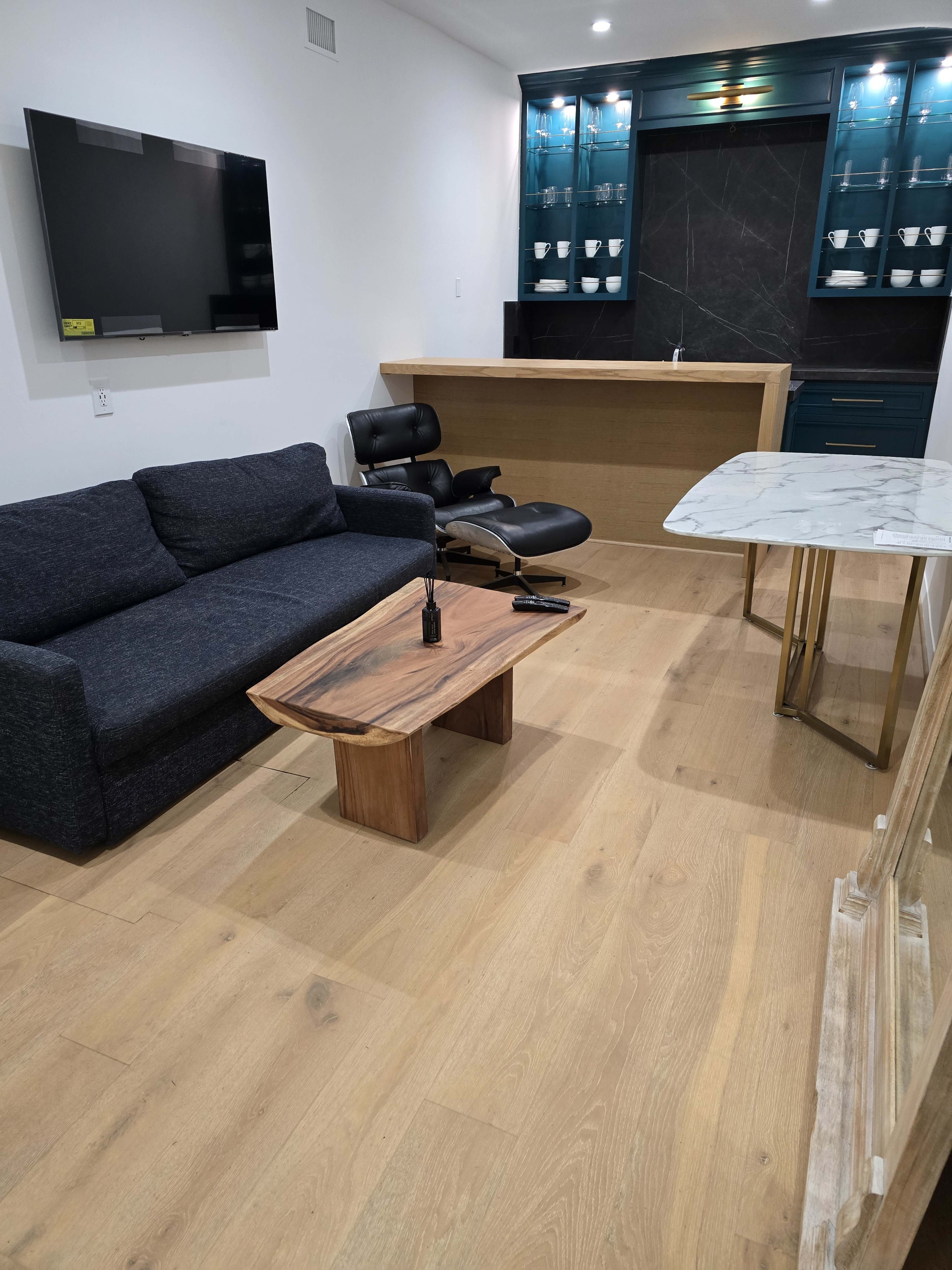 The image shows a modern living space with a dark sofa, a wooden coffee table, and a sleek kitchen bar area featuring glass cabinets and a marble-topped table.