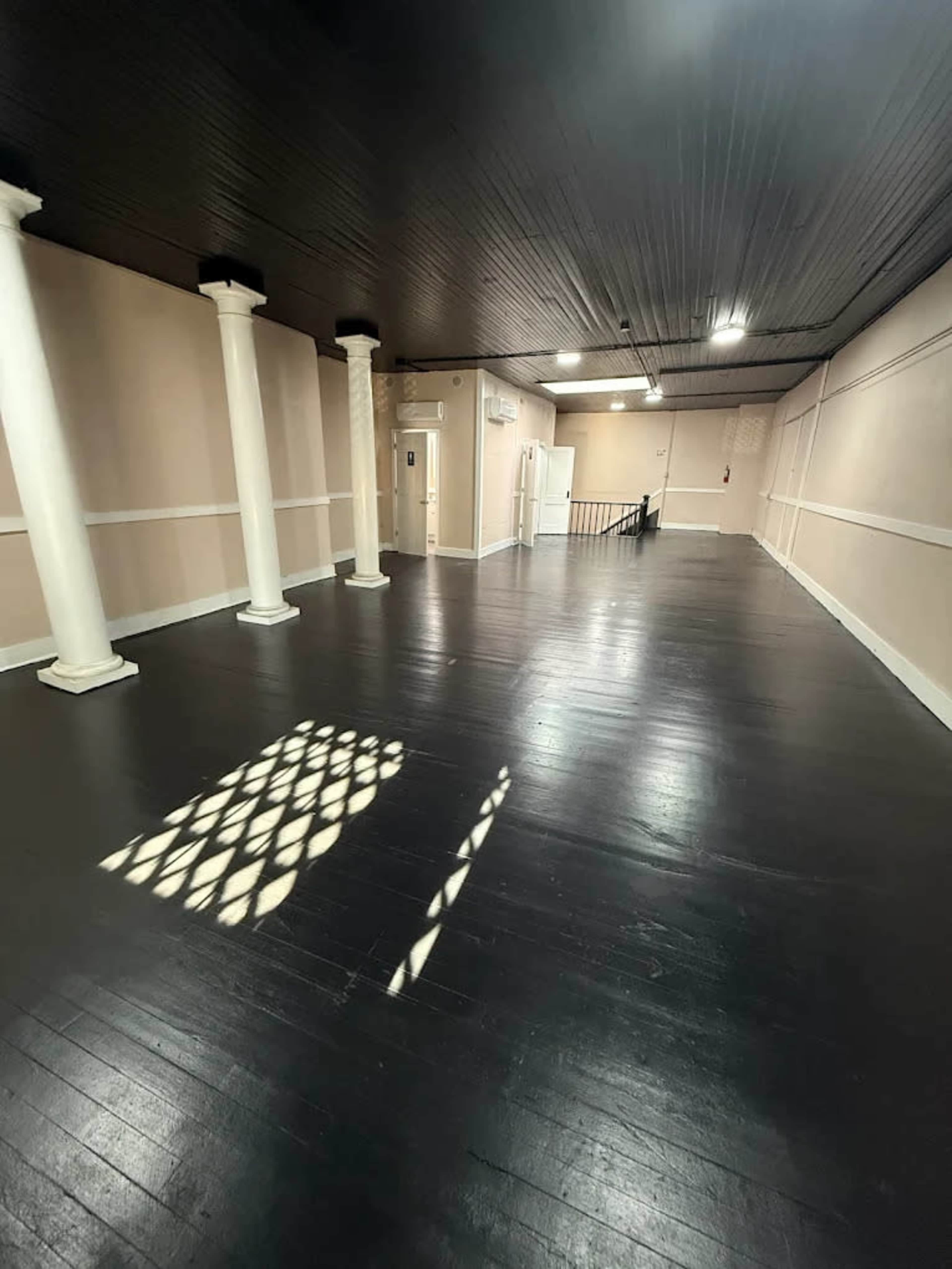 The image shows a spacious, empty room with black wooden flooring, white columns, and a staircase leading to another area.