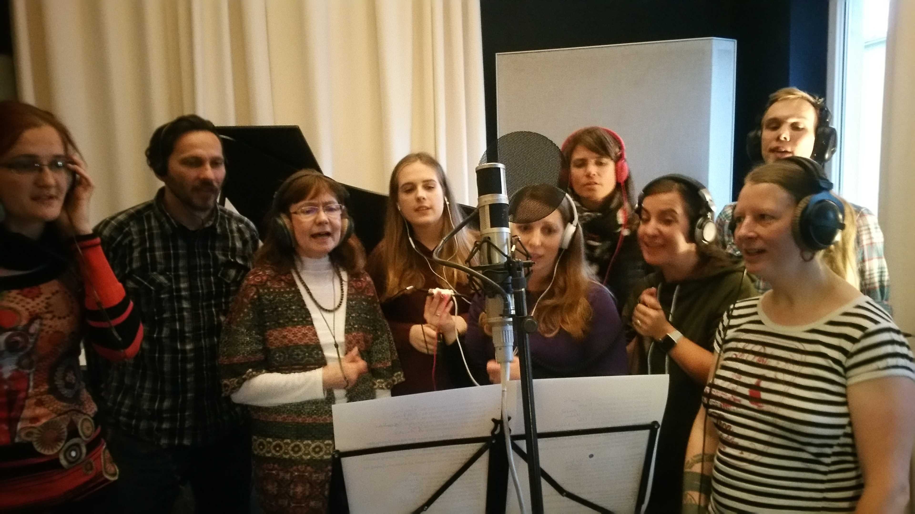 A group of eight people wearing headphones stands in a recording studio, gathered around a microphone while singing.