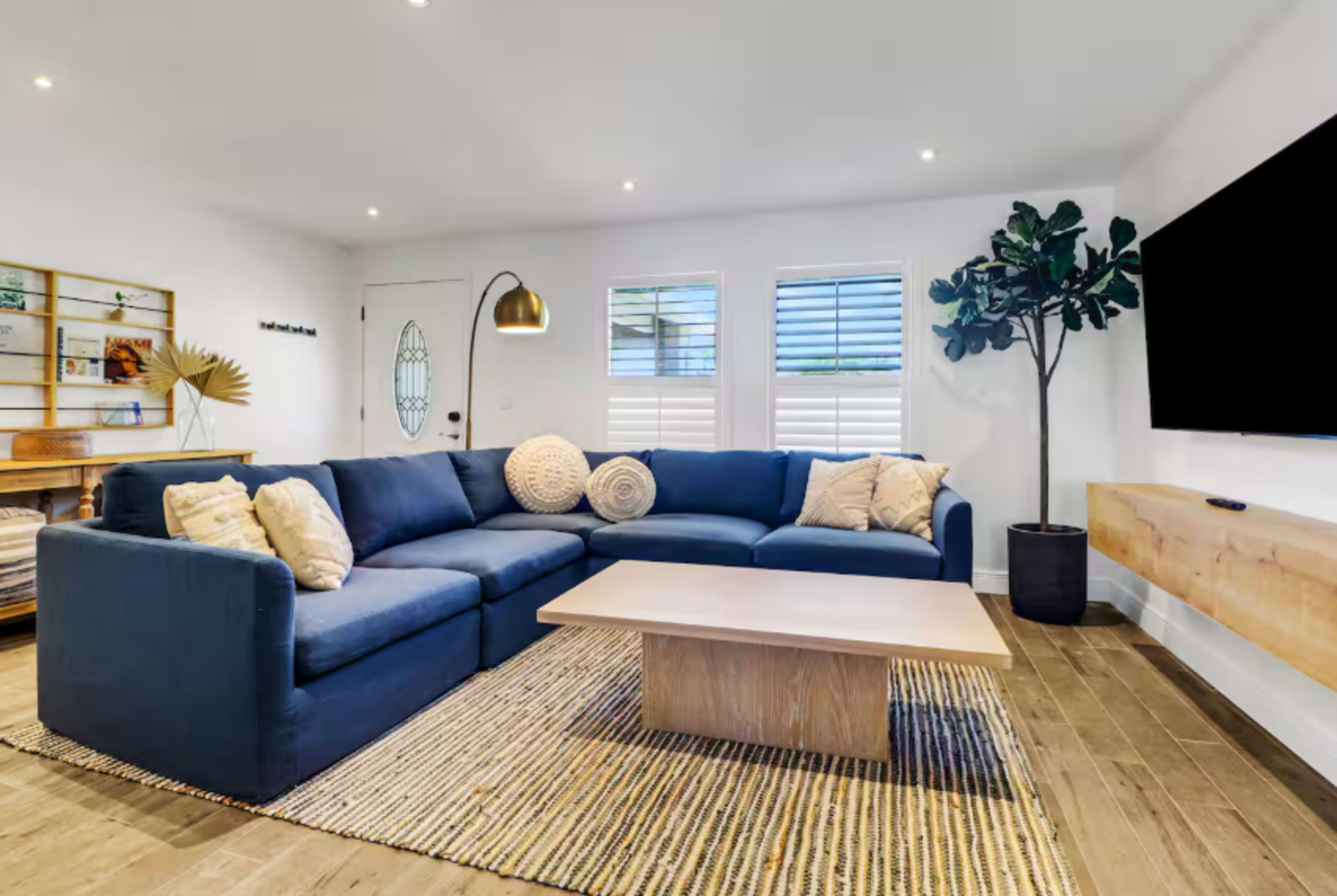 A modern living room features a large blue sectional sofa, a wooden coffee table, and a decorative plant, with light-colored walls and natural light coming through the windows.