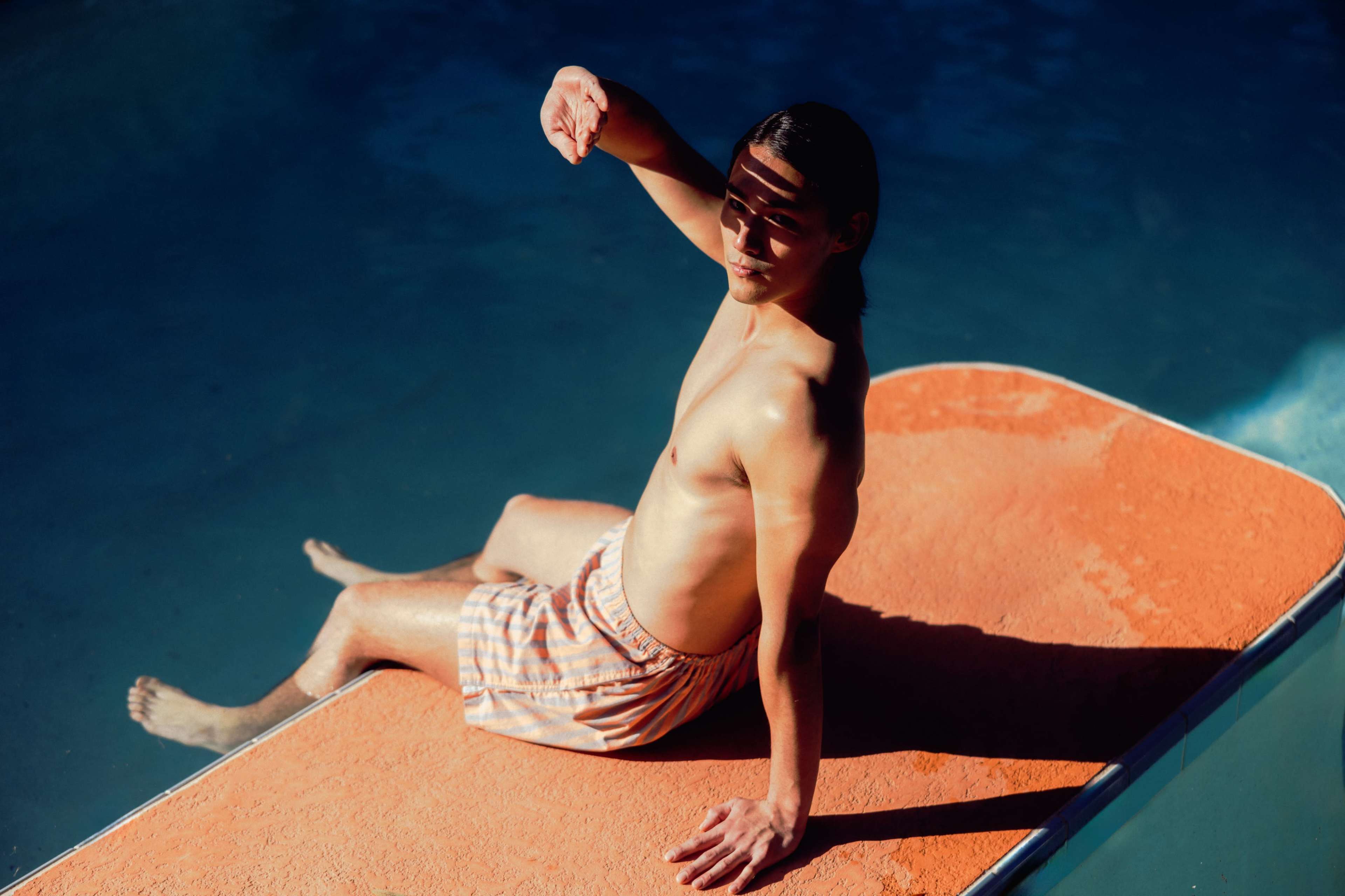 A young man sits on the edge of a pool wearing striped swim shorts, with shadows casting on his body from nearby structures.