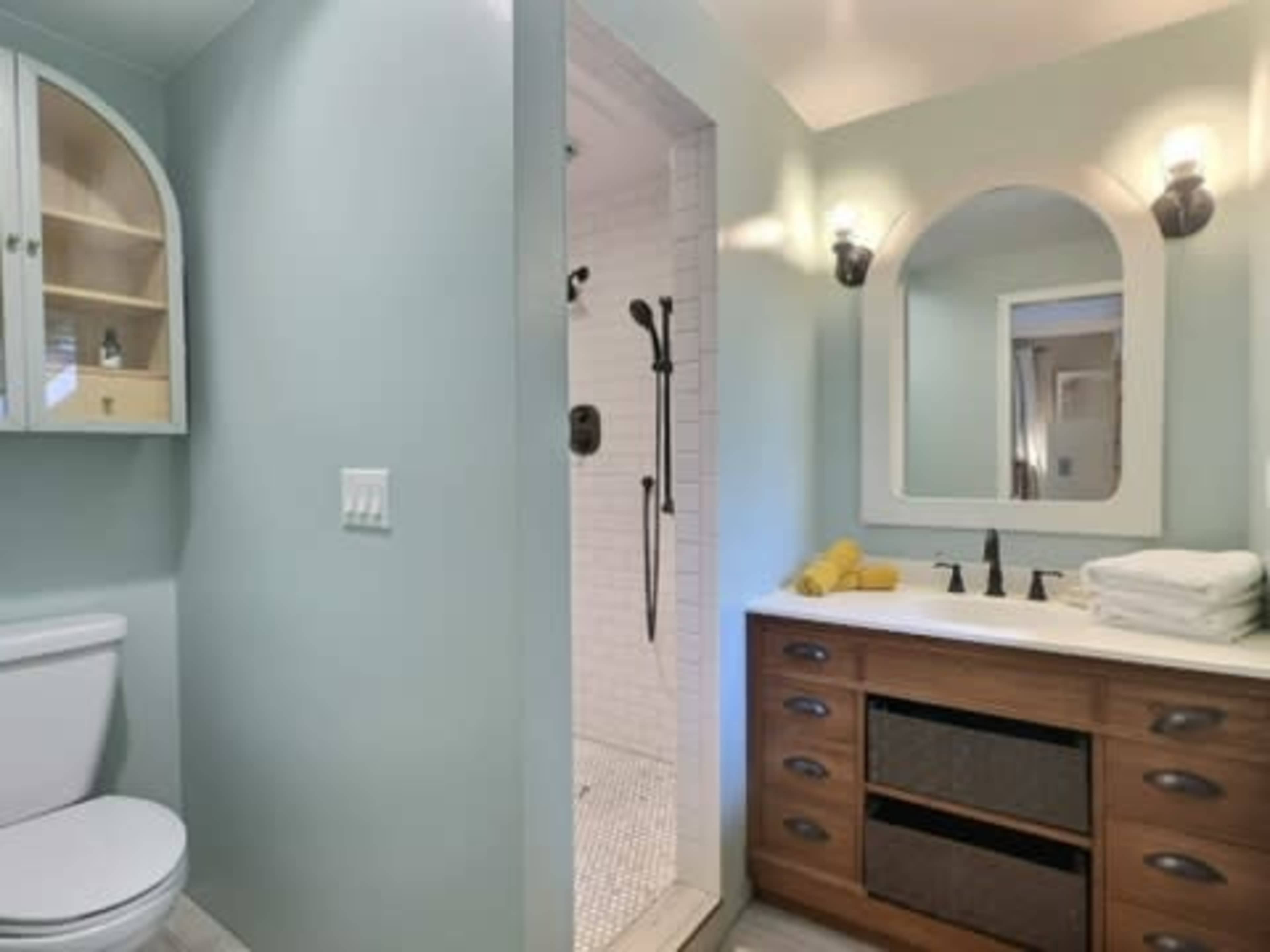 The image shows a bathroom featuring a shower area, a wooden vanity with storage, white towels, and a toilet.