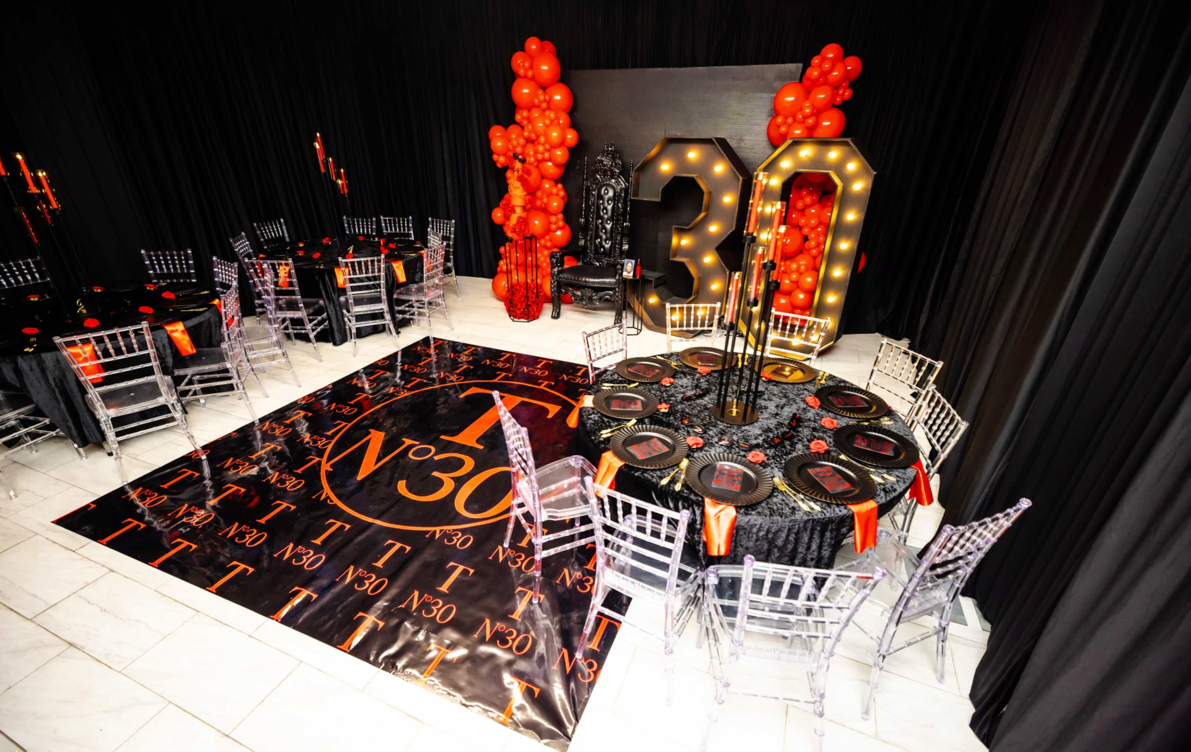 The image shows a decorated event space featuring tables with black linens, illuminated "30" signage, and red balloon arrangements.