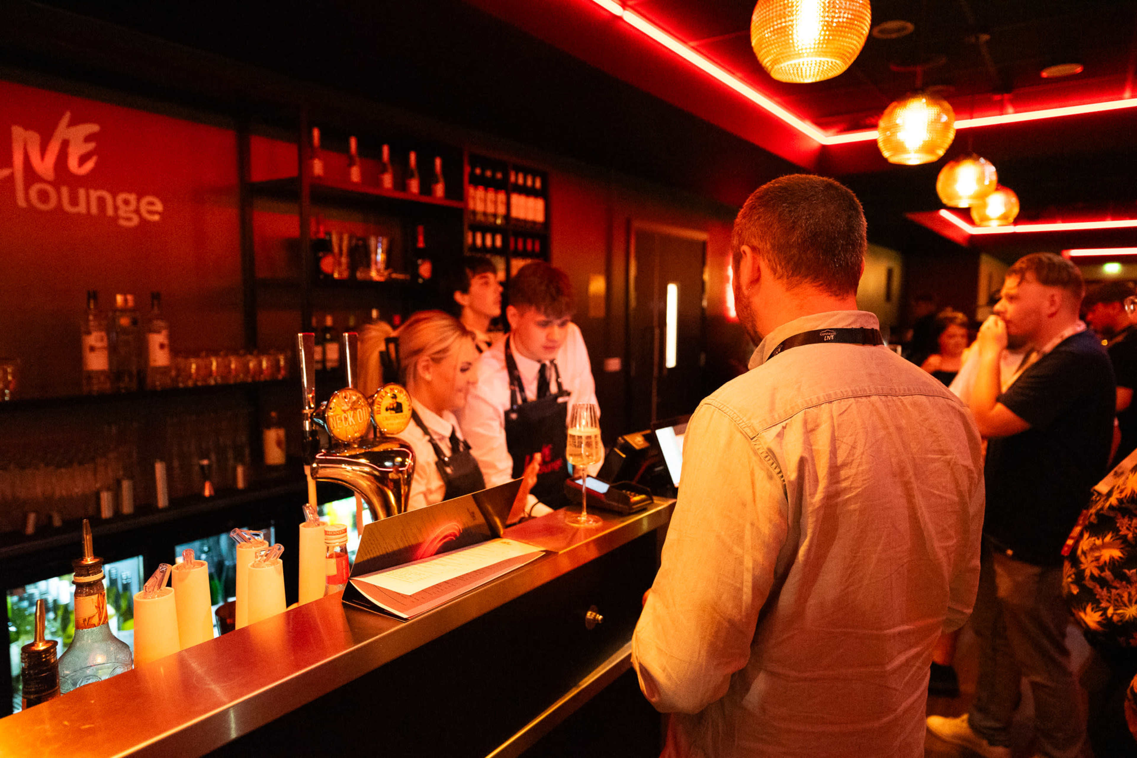 A bartender serves drinks at a well-lit bar while patrons gather around in a lounge setting.