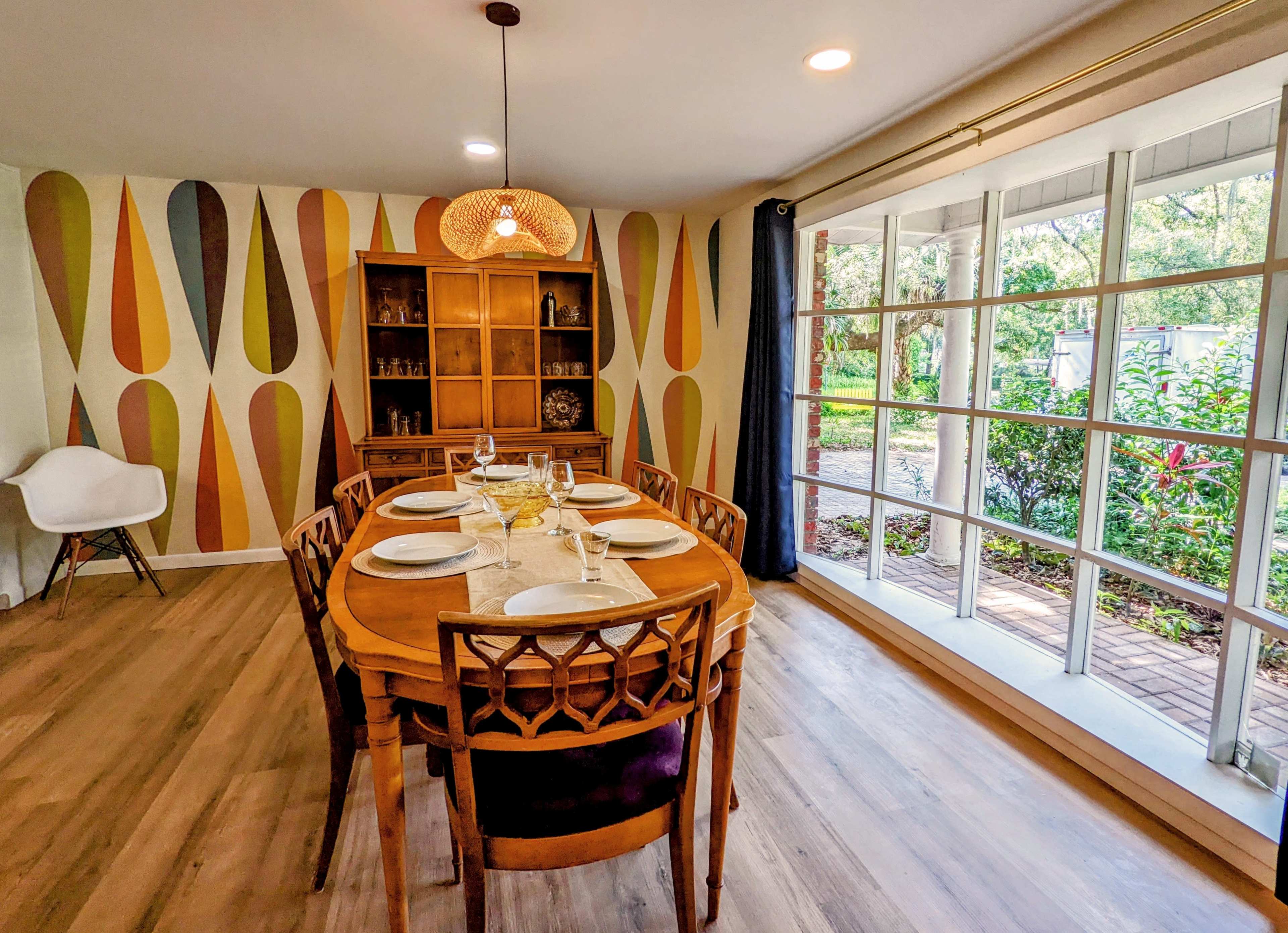 A dining room features a round wooden table set for six, surrounded by chairs, with a patterned wallpaper and large windows showing greenery outside.