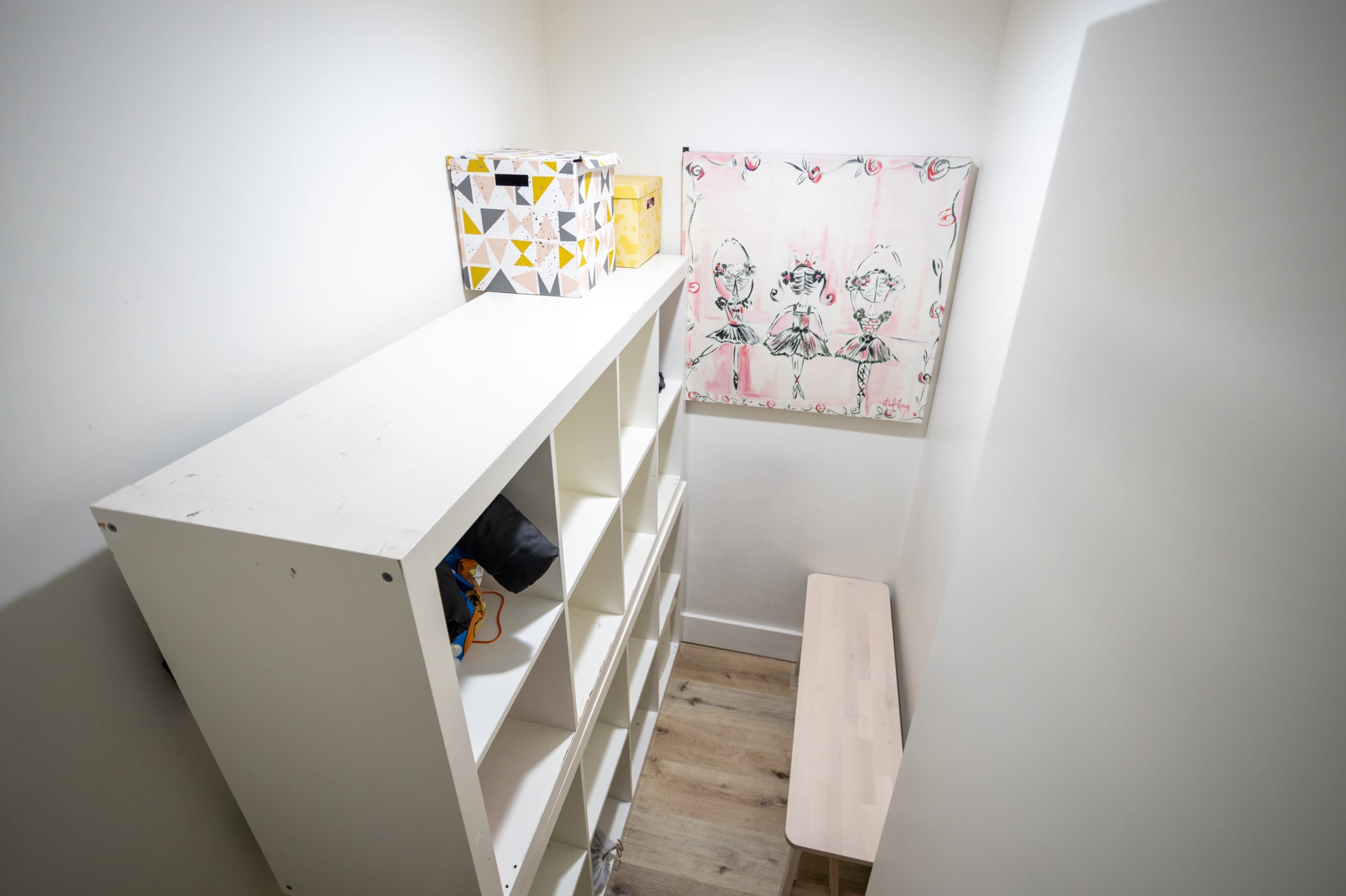 A small, organized room features a white shelving unit and a light wooden bench against a wall decorated with a colorful artwork.