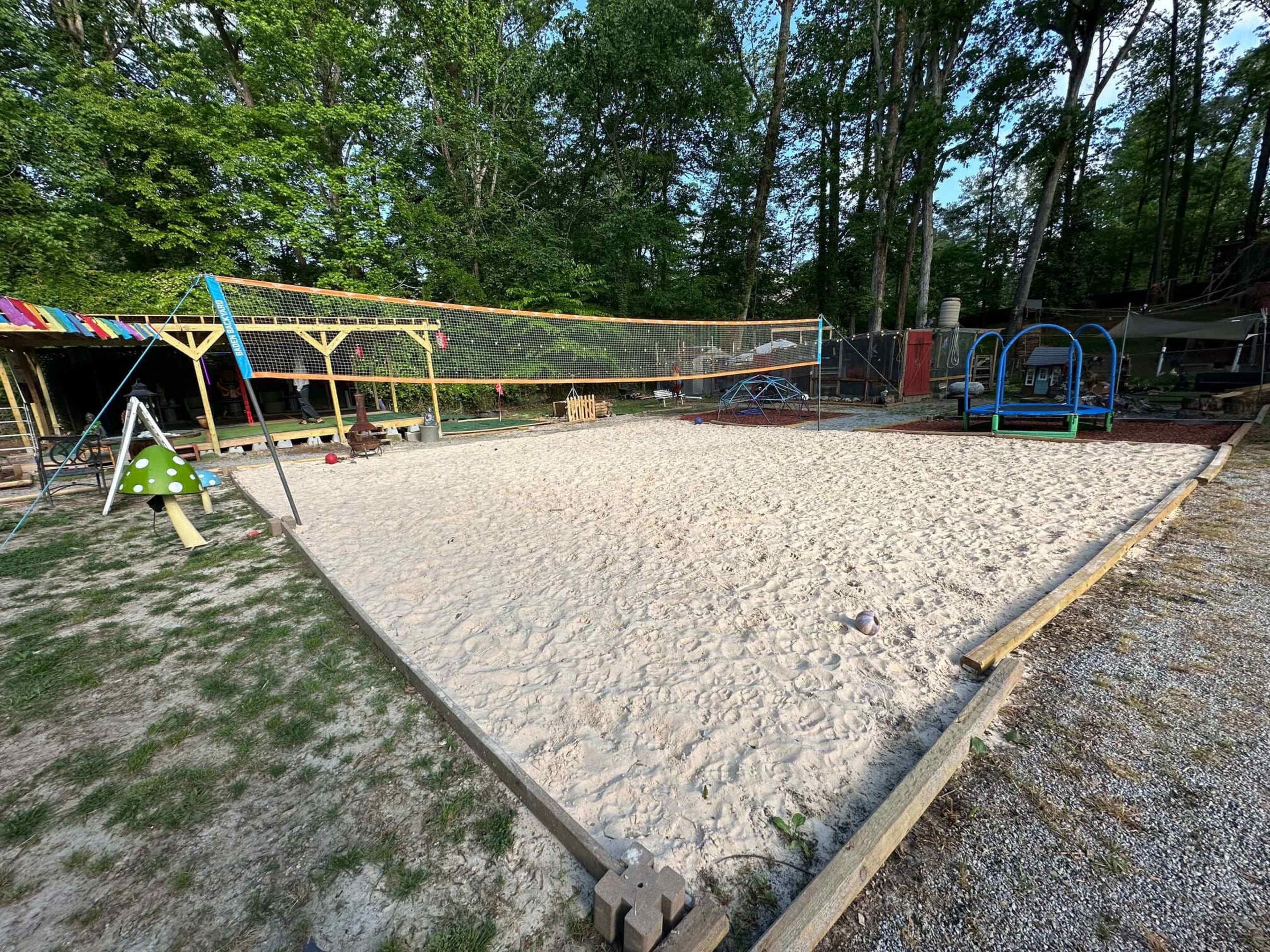 The image shows a sandy area that appears to be a volleyball court, surrounded by greenery, with playground equipment and a fence in the background.