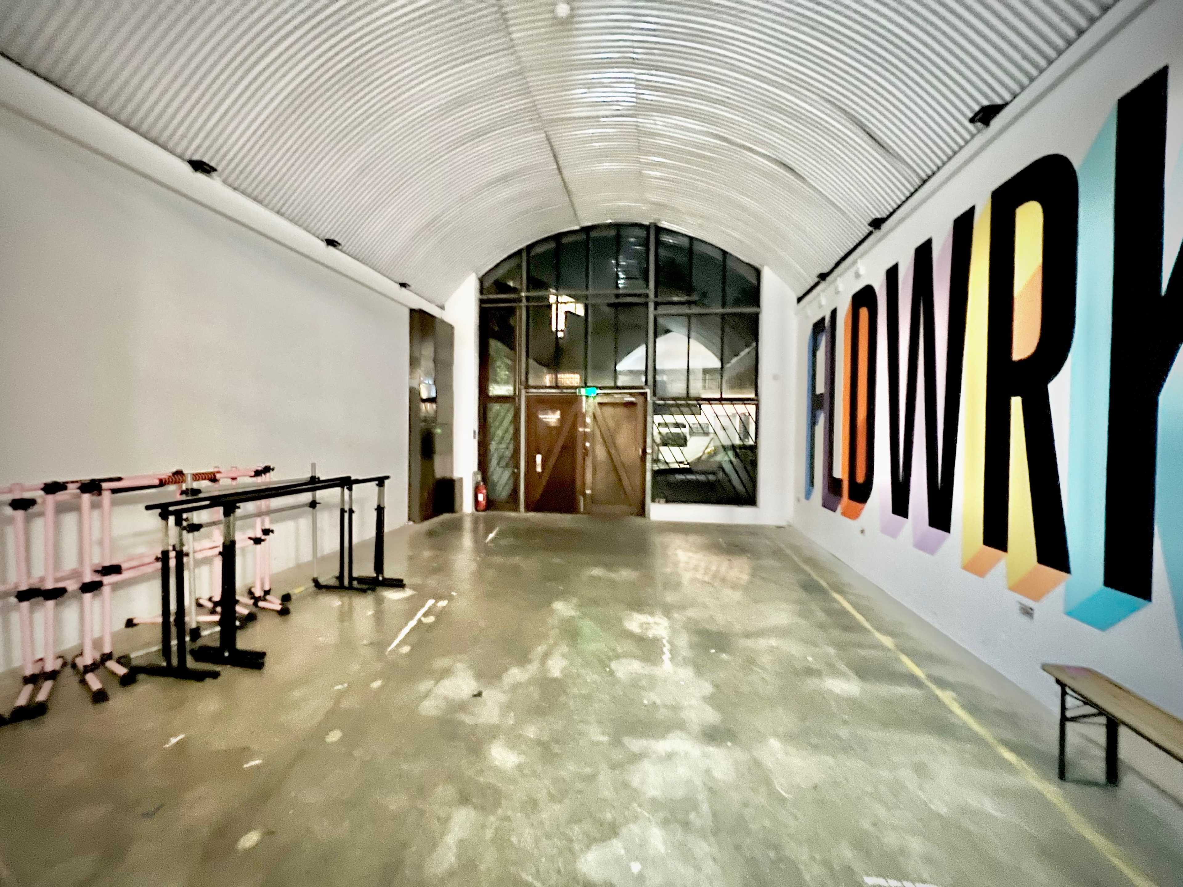 A spacious, minimalist interior with a high arched ceiling, a large window at one end, and a colorful mural reading "FLDWRK" on the wall, alongside several standing exercise bars lined up against the wall.