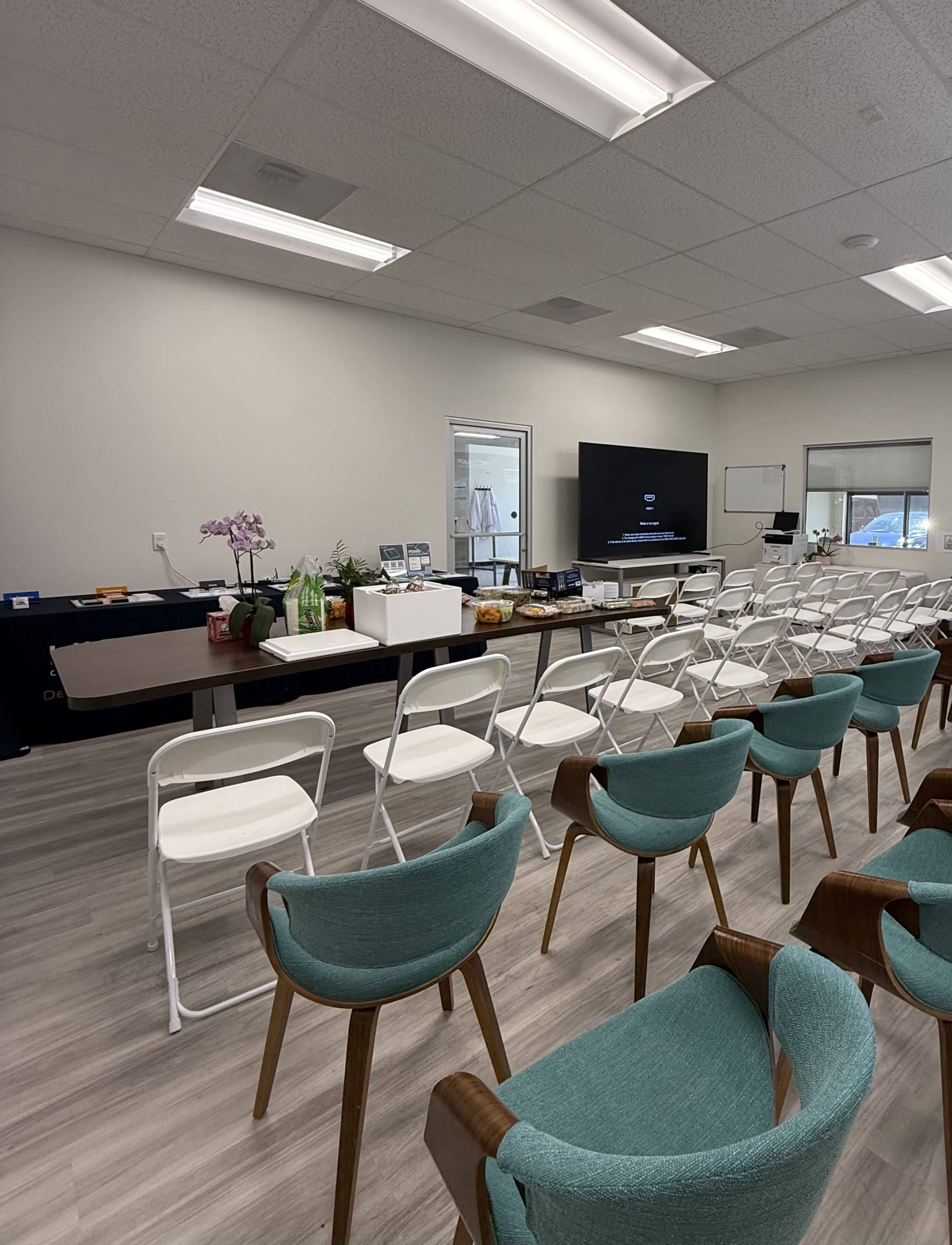 The image shows a modern meeting room with rows of white chairs facing a large screen, along with a table displaying refreshments and a potted orchid.