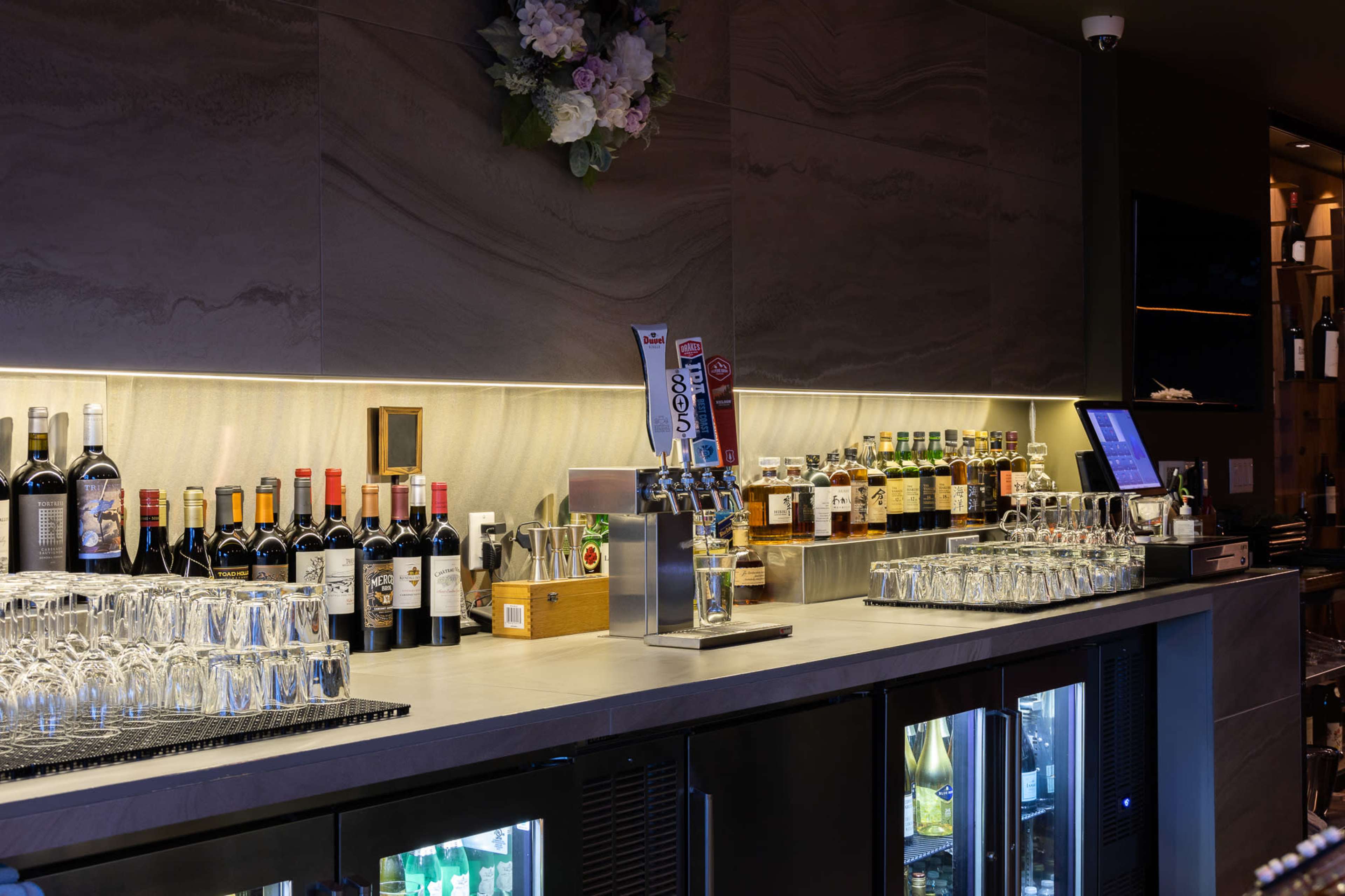 The image shows a well-stocked bar counter featuring various bottles of wine and spirits, alongside glassware and beer taps under soft lighting.