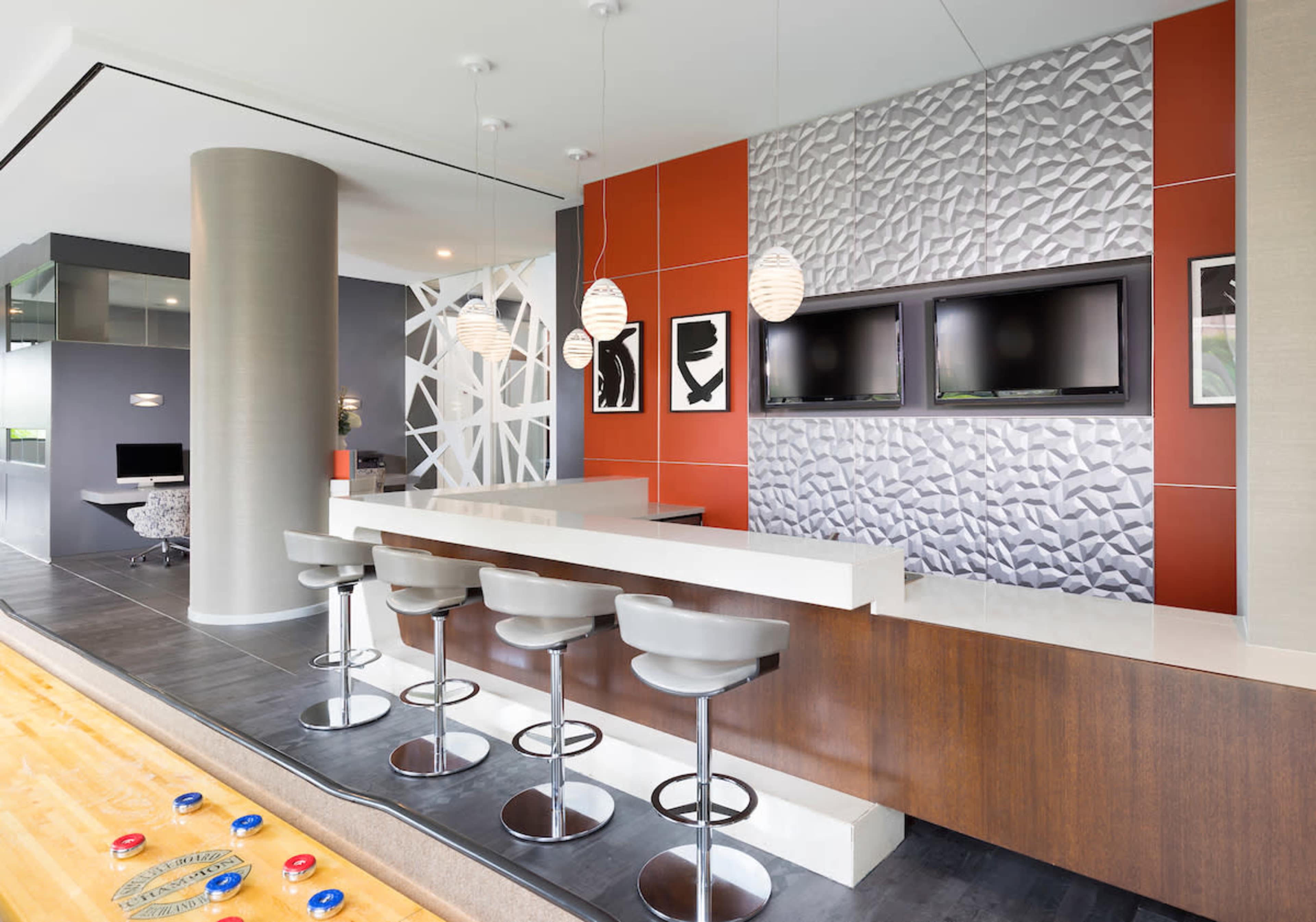 A modern kitchen with a sleek bar area featuring four stools, a textured wall, and two television sets mounted above a counter.