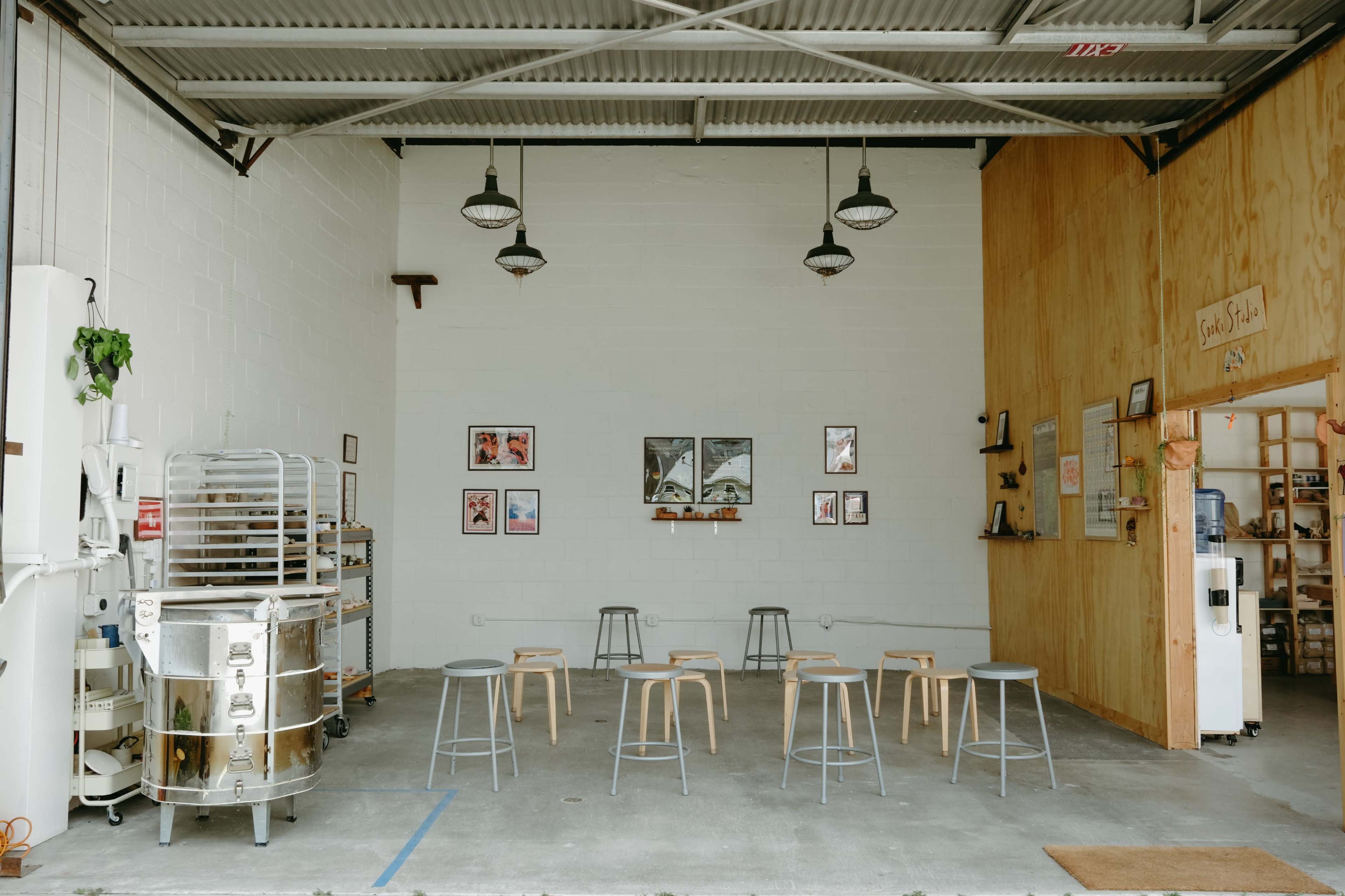 A minimalist cafe interior with wooden stools arranged around a concrete floor beneath industrial light fixtures and framed artwork on the walls.