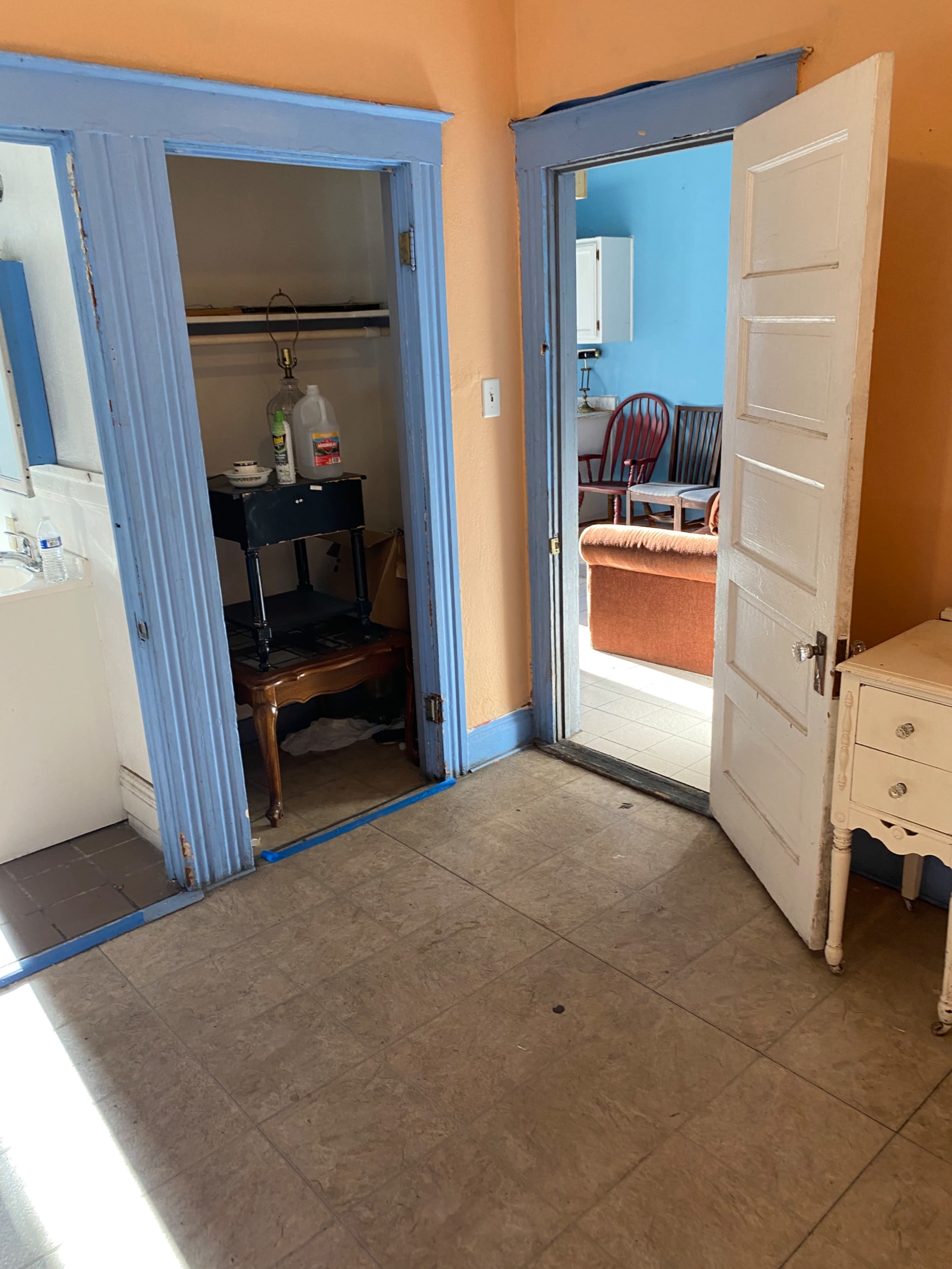 A room with two doorways leading to adjacent spaces, featuring a small table and a wardrobe in one area, while a sofa and chair are visible in the other room.