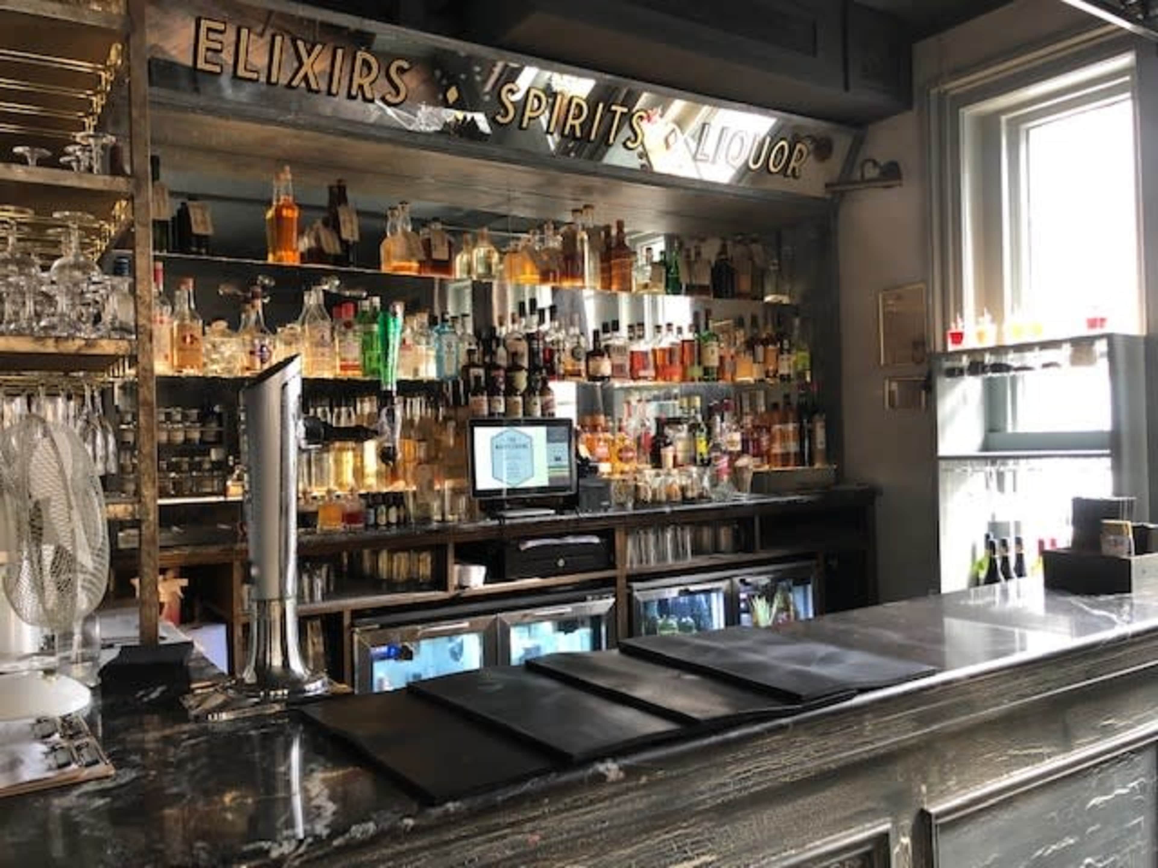 The image shows a bar with a well-stocked shelf of various liquor bottles, a display screen, and several menus laid out on the counter.
