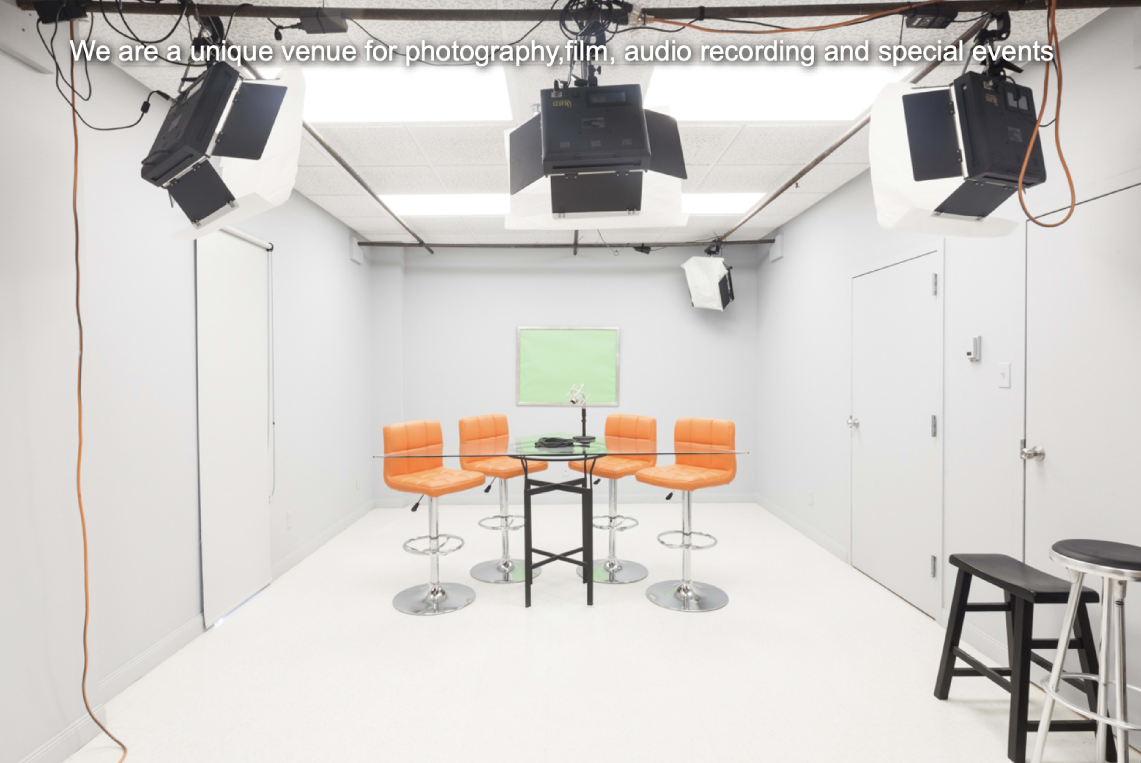 A bright, minimal studio space features a circular table with black legs surrounded by four orange bar stools, and is equipped with multiple overhead lights.