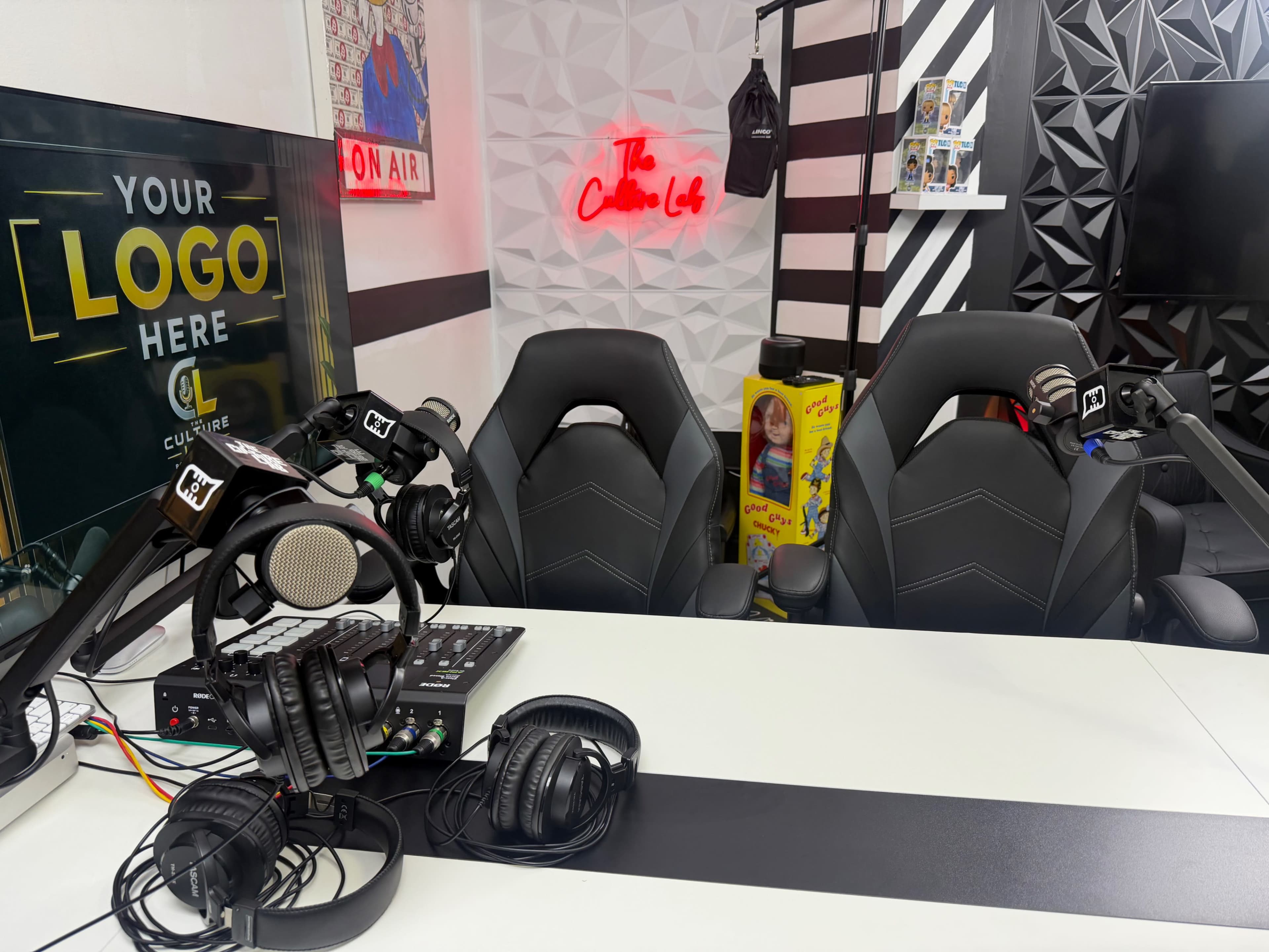 The image shows a modern podcast studio setup with two empty black chairs, microphones, headphones, and a mixing console on a white table, along with a neon sign in the background.