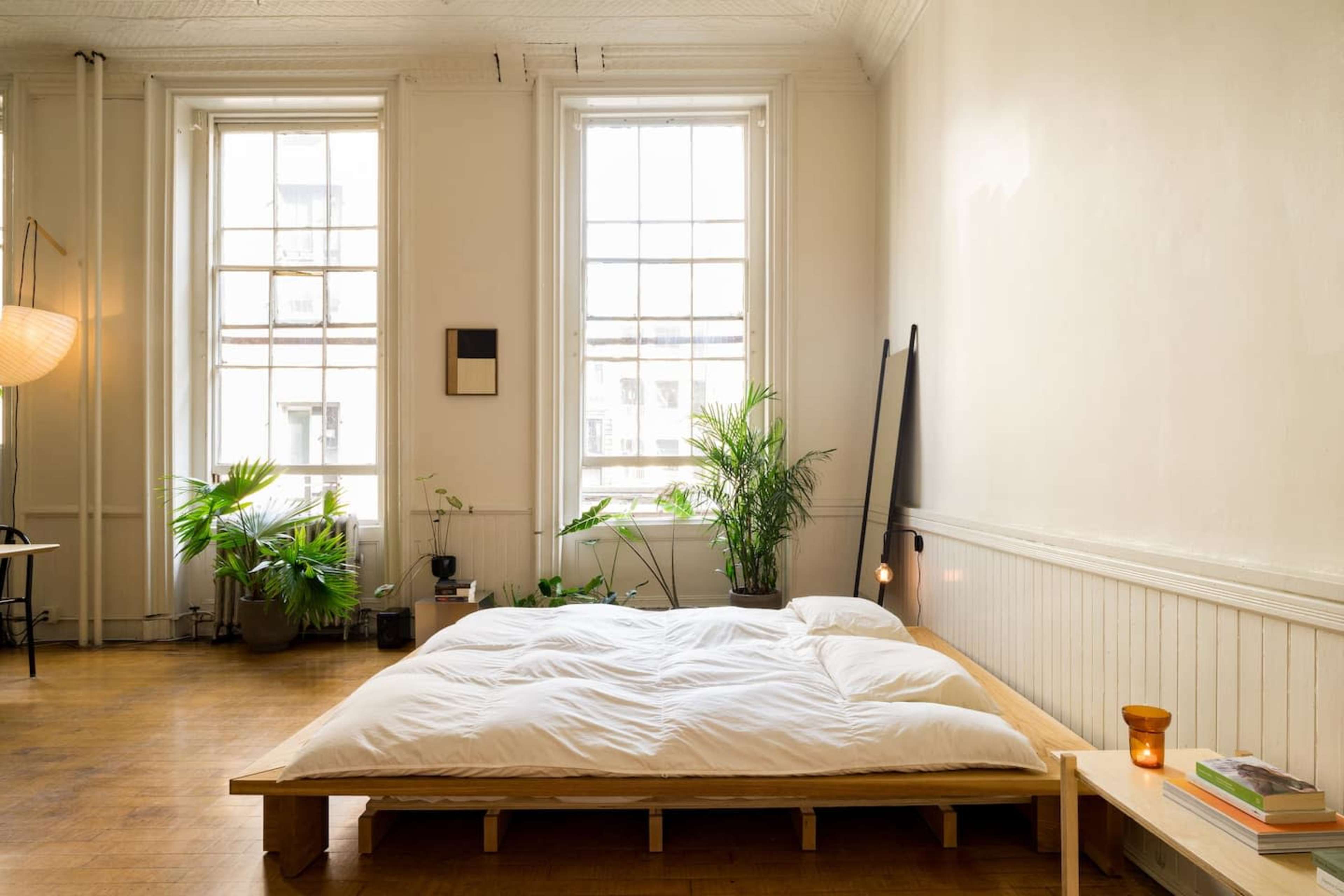 A minimalistic bedroom features a low wooden bed with a white comforter, large windows letting in natural light, and several potted plants.